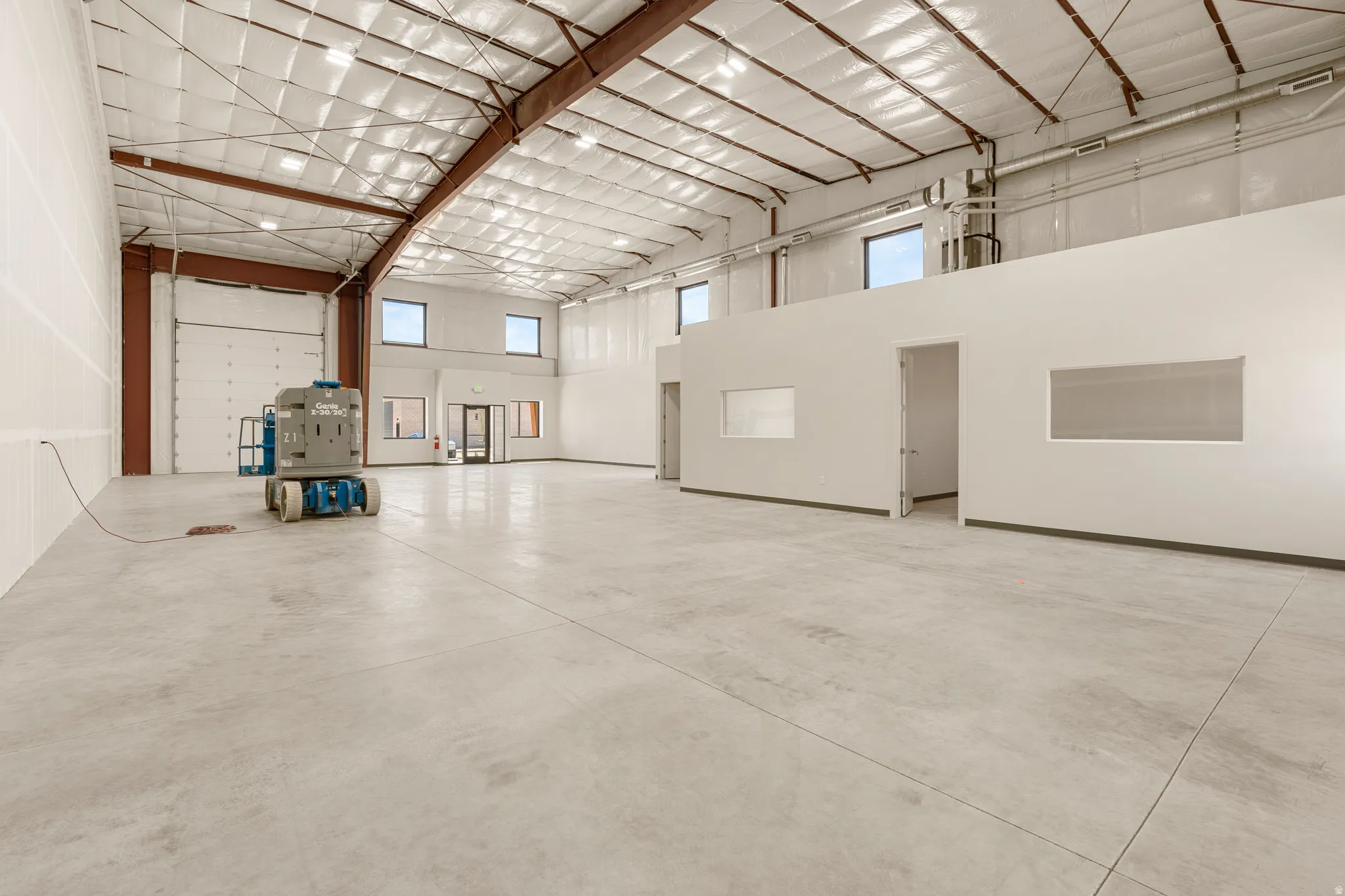 Spacious commercial interior featuring concrete flooring, a high exposed ceiling with metal beams, multiple windows providing natural light, and an integrated office area with interior windows