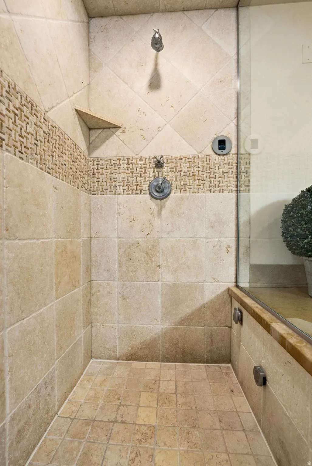 Shower enclosure featuring natural stone-look tile, a decorative mosaic tile band, a built-in corner shelf, and a glass partition