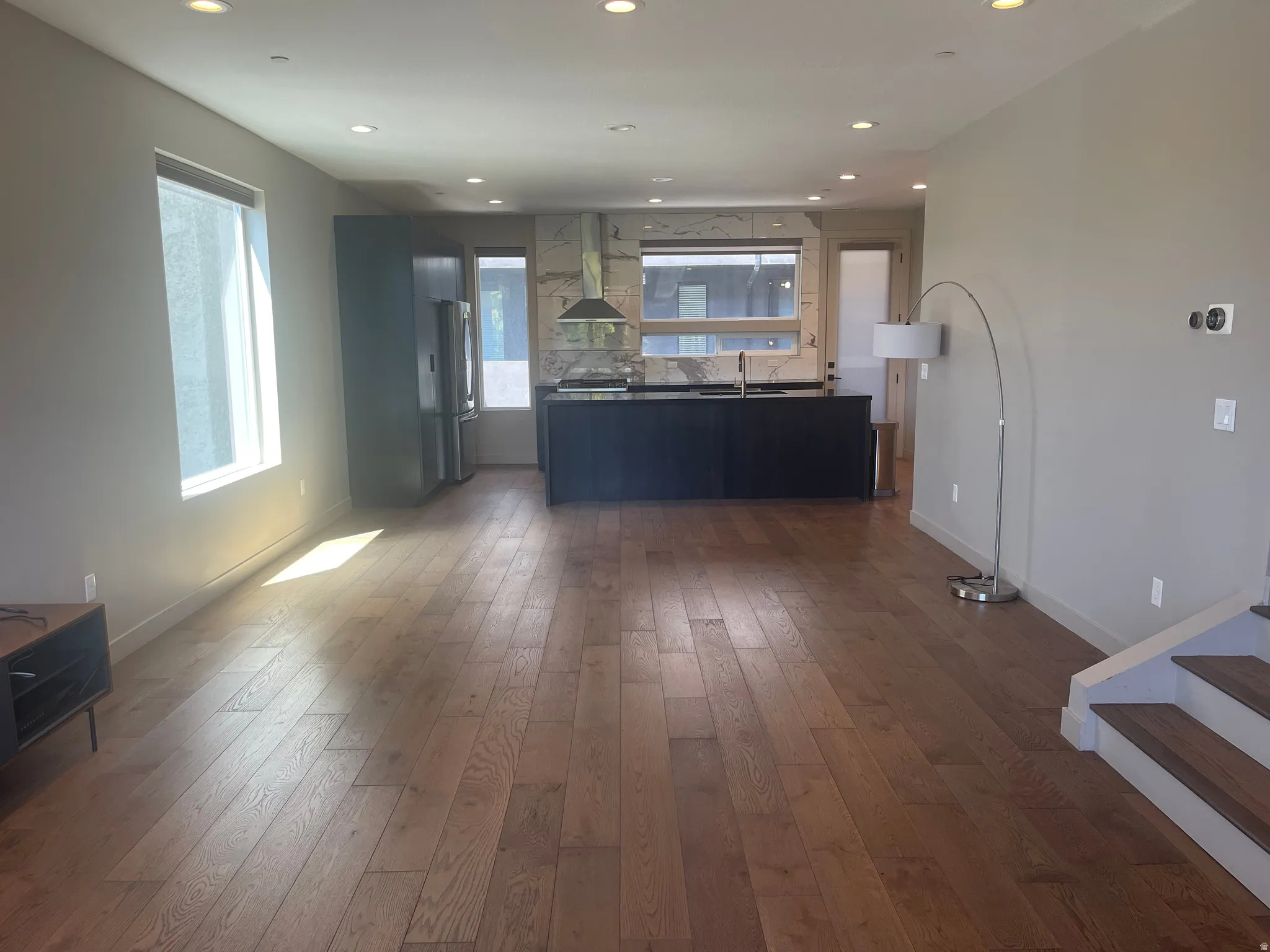 Open-concept living area featuring wood-finish flooring, recessed lighting, and a kitchen with an island, dark cabinetry, stainless steel appliances, and a full-height backsplash