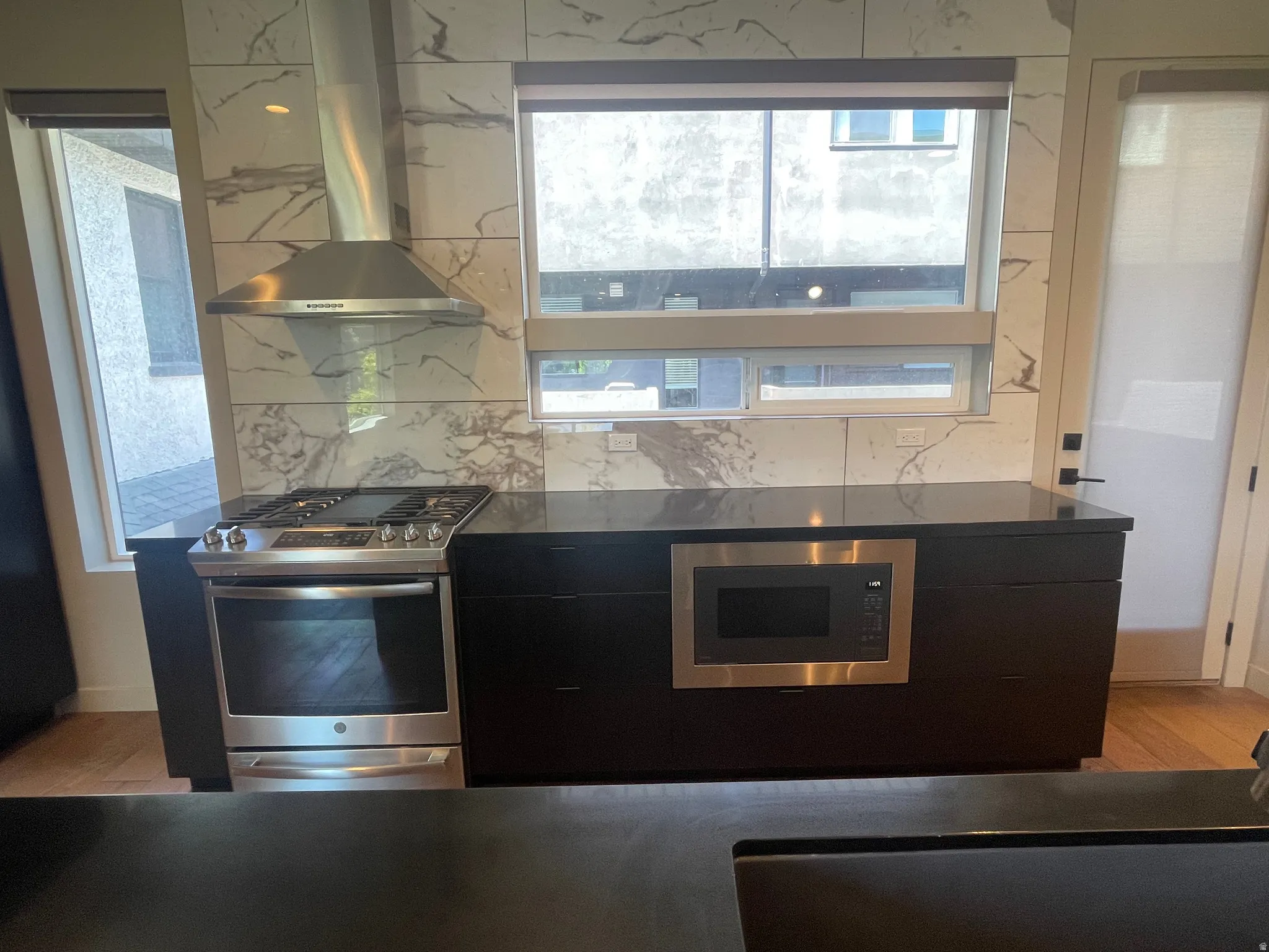 Kitchen featuring a stainless steel range with a ventilation hood, built-in microwave, dark cabinetry, dark countertops, and large format marble-look tile backsplash