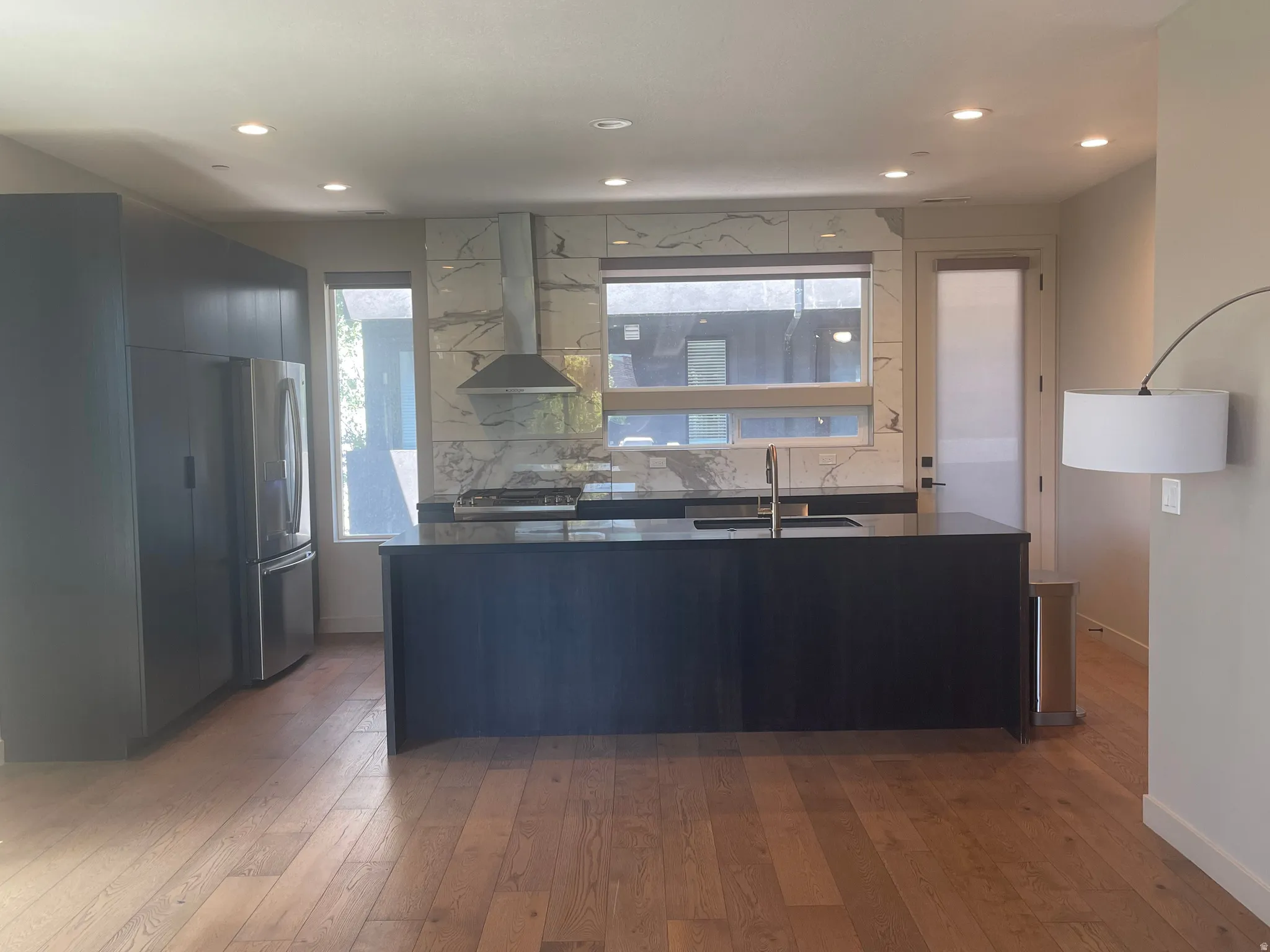 Modern kitchen featuring sleek dark cabinetry, a large center island with an integrated sink, a stainless steel range hood, and a full-height marble-look backsplash
