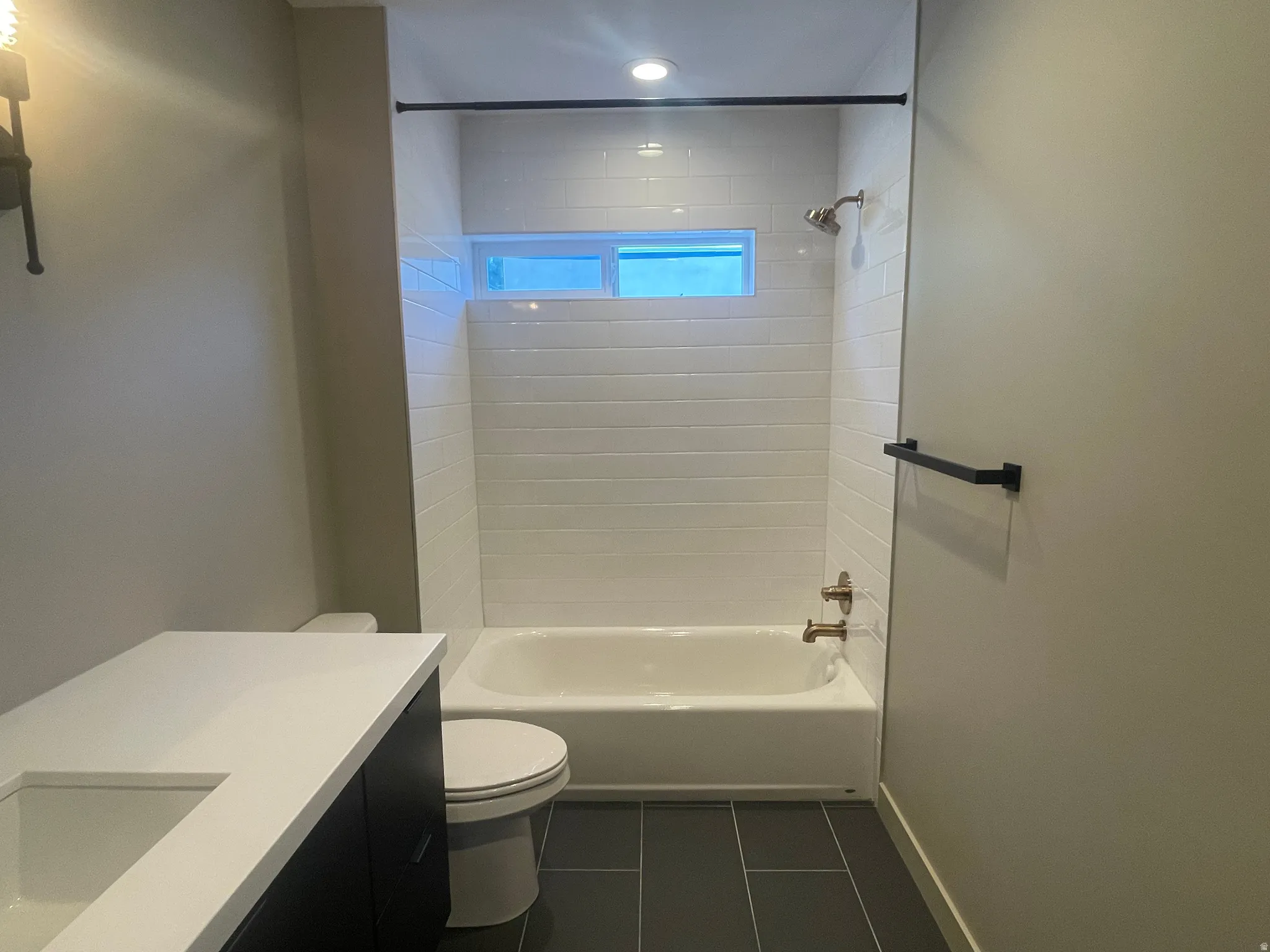 Full bathroom featuring a vanity with an integrated sink and white countertop, white subway tile shower surround, shower/tub combination with bronze fixtures, and rectangular dark tile flooring