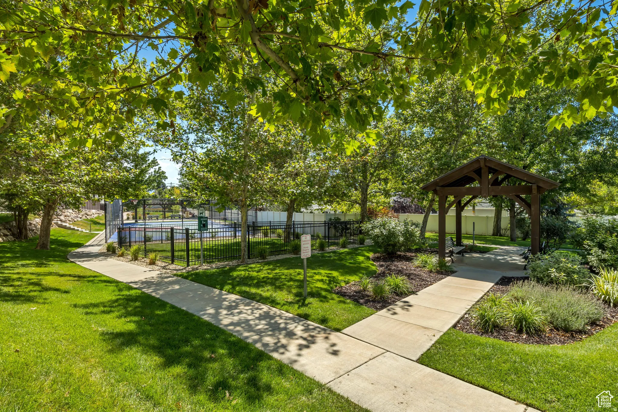 Outdoor amenity space featuring a paved pathway, expansive lawn, mature shade trees, and a wooden gazebo