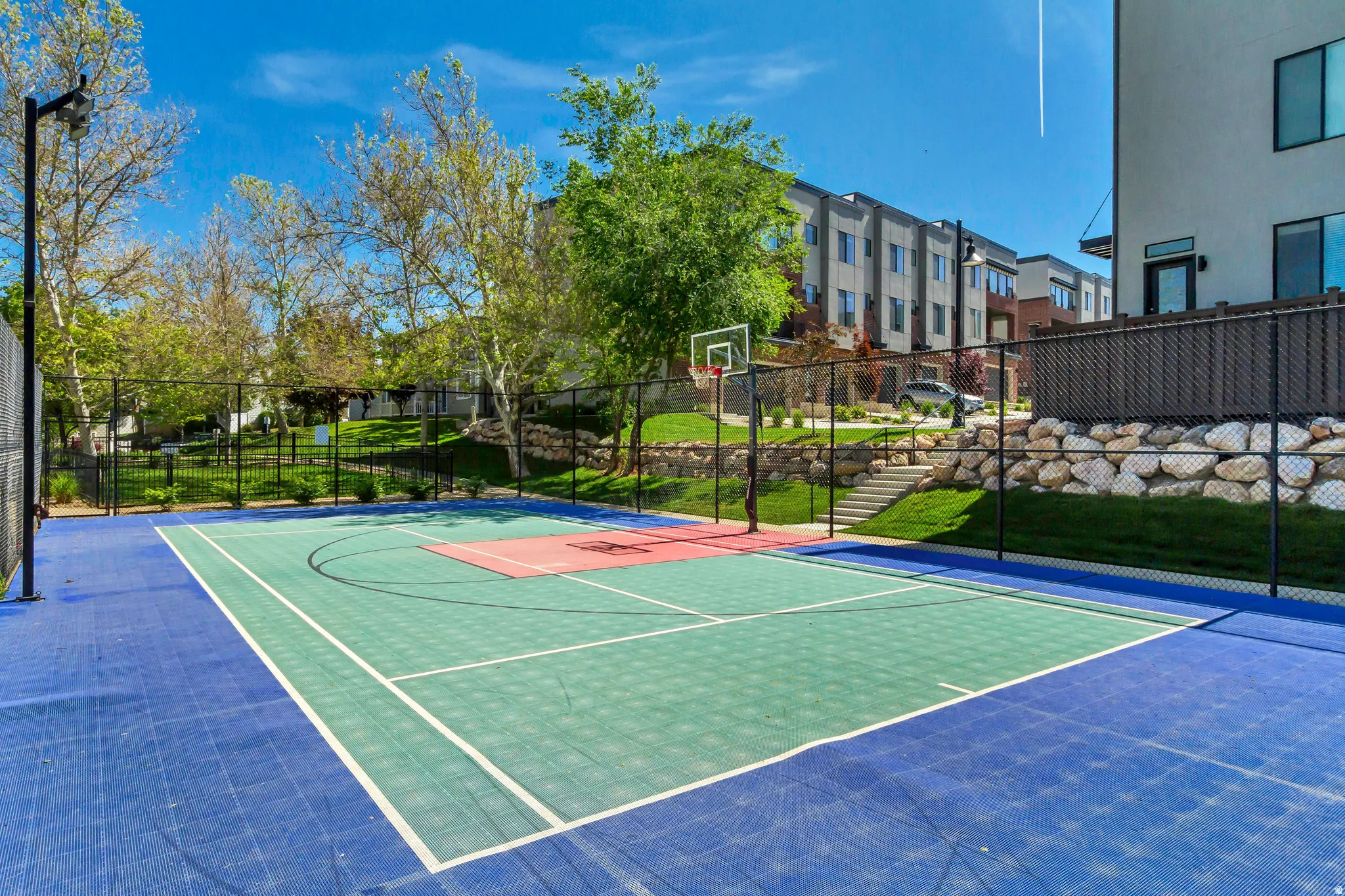 Multi-color outdoor sports court with a basketball hoop, surrounded by black chain-link fencing