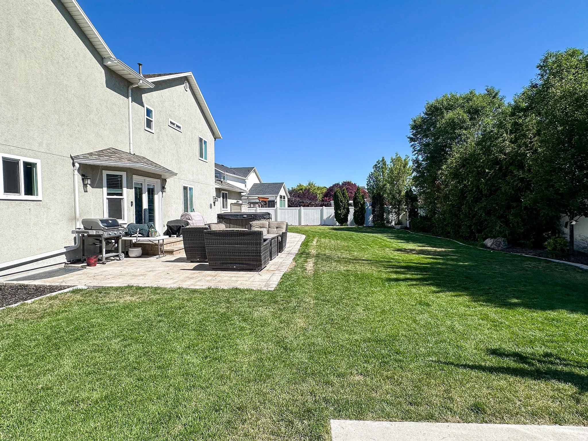Expansive backyard featuring a large paver patio, a wide lawn area, mature perimeter trees, and a white privacy fence