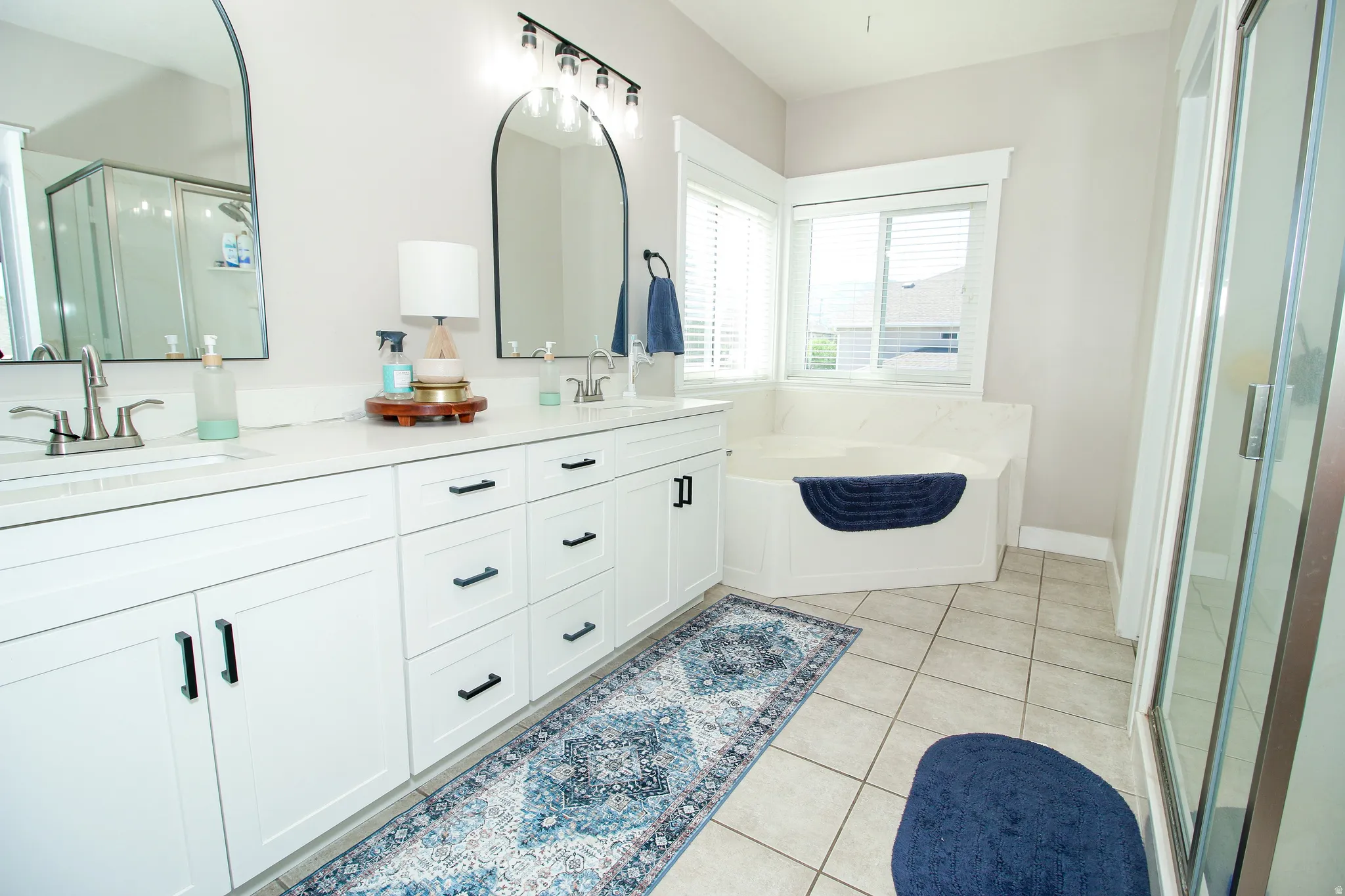 Bright bathroom featuring a double vanity with white cabinetry and black hardware, dual arched mirrors, a corner soaking tub, and a glass-enclosed shower