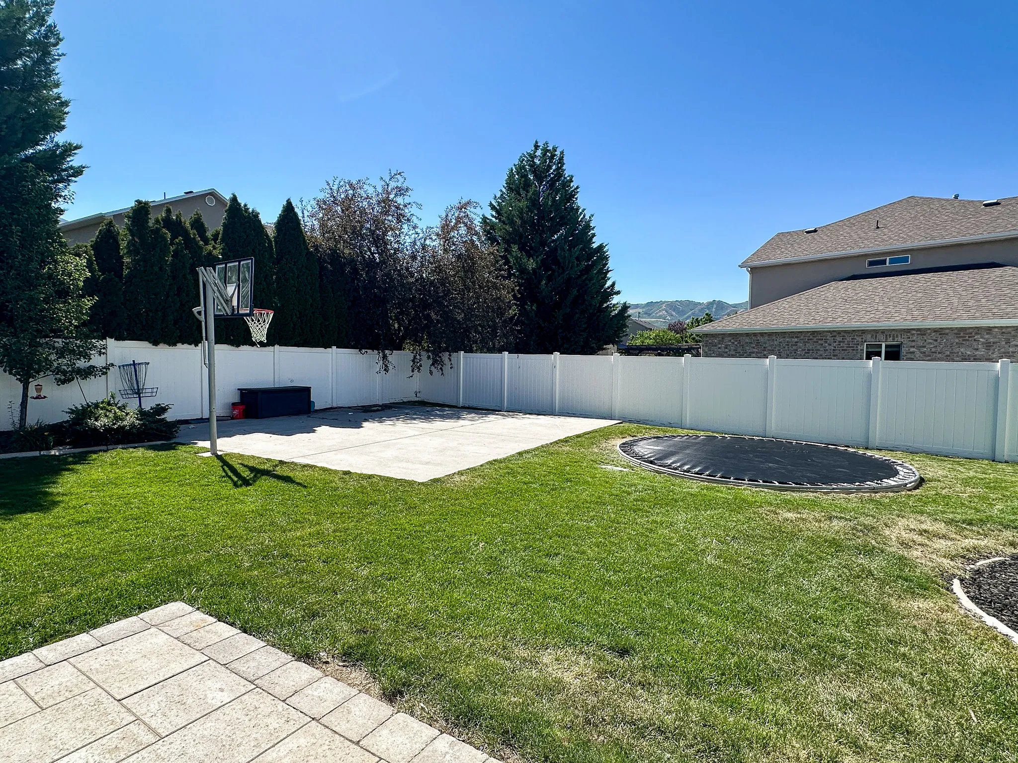 Expansive backyard featuring a lush lawn, white vinyl fencing, a concrete pad, a built-in trampoline, and mature landscaping