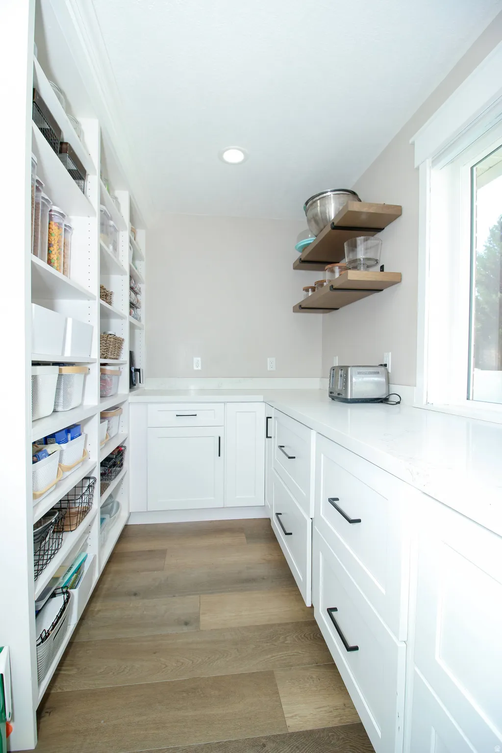 Kitchen pantry featuring extensive built-in shelving, white cabinetry with black hardware, light-toned wood-finish flooring, white countertops, and a window providing natural light