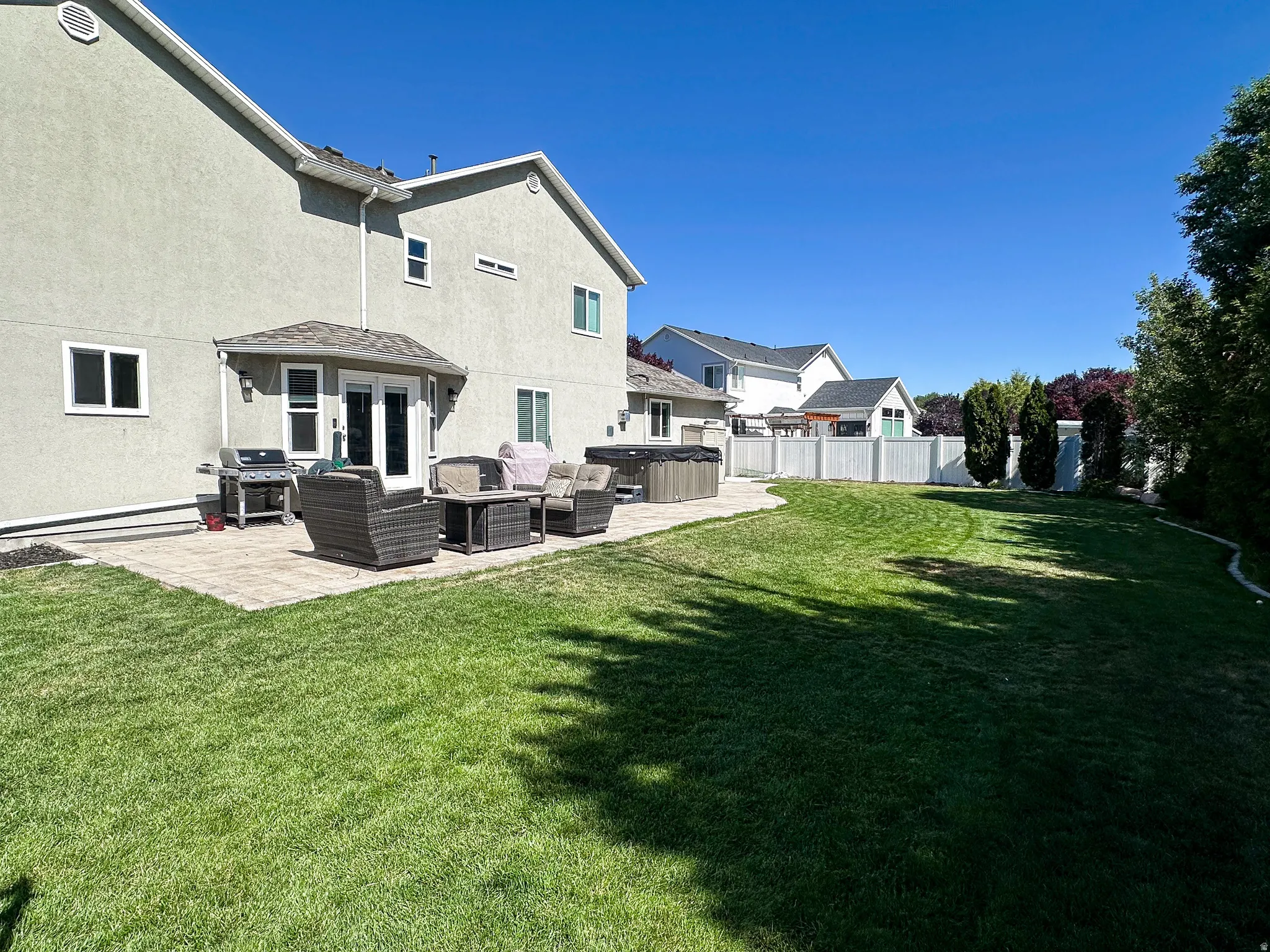 Spacious backyard featuring a large paver patio, lush green lawn, white privacy fencing, and mature perimeter trees