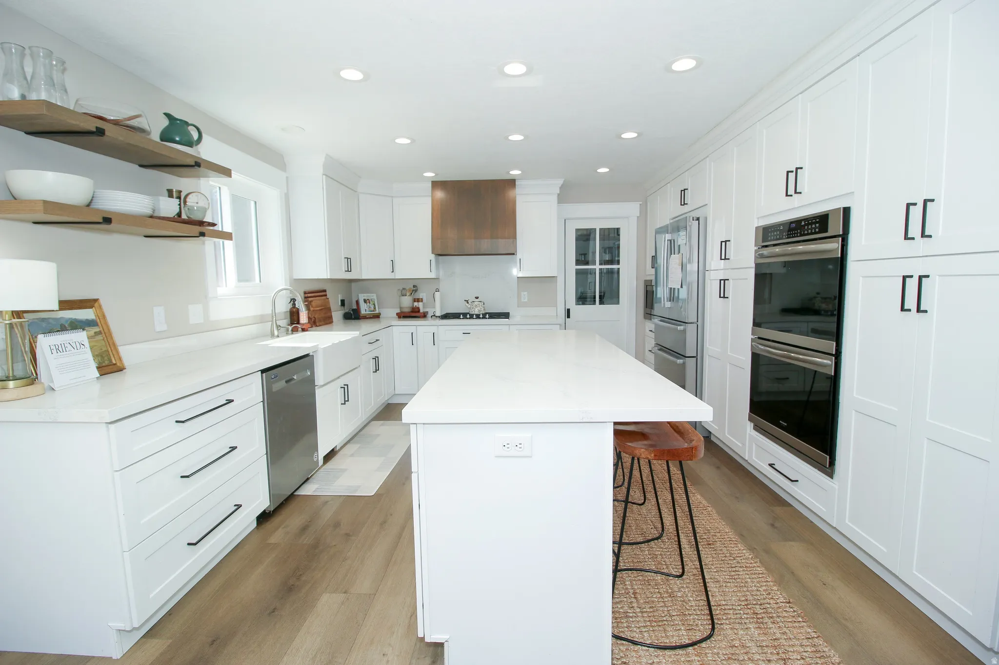 Bright kitchen featuring a substantial center island with seating, white shaker cabinetry, stainless steel appliances, wood-finish flooring, and open shelving