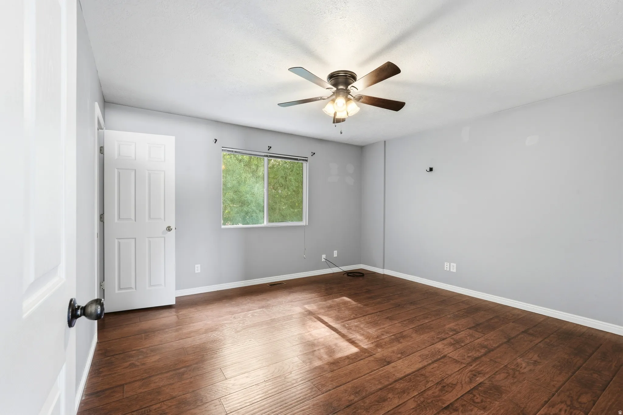 Spacious room featuring wood-finish flooring, a ceiling fan with integrated lighting, and a window providing natural light