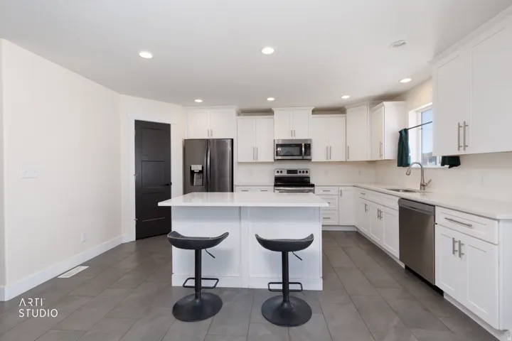 Modern kitchen featuring a central island with white paneling and a light-toned countertop, white cabinetry with silver-toned hardware, stainless steel appliances, recessed ceiling lighting, and dark tile flooring