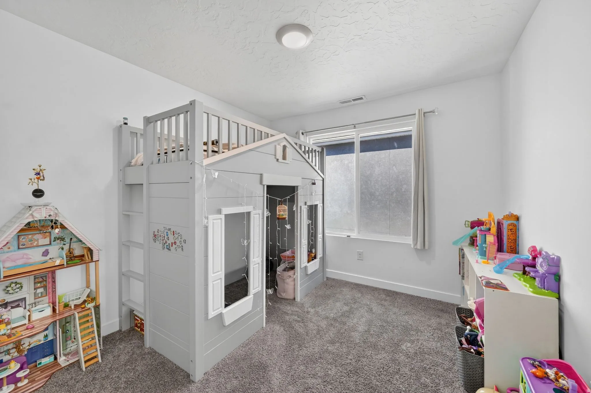 White walls complement the grey carpet flooring and a large window with white trim and grey curtains