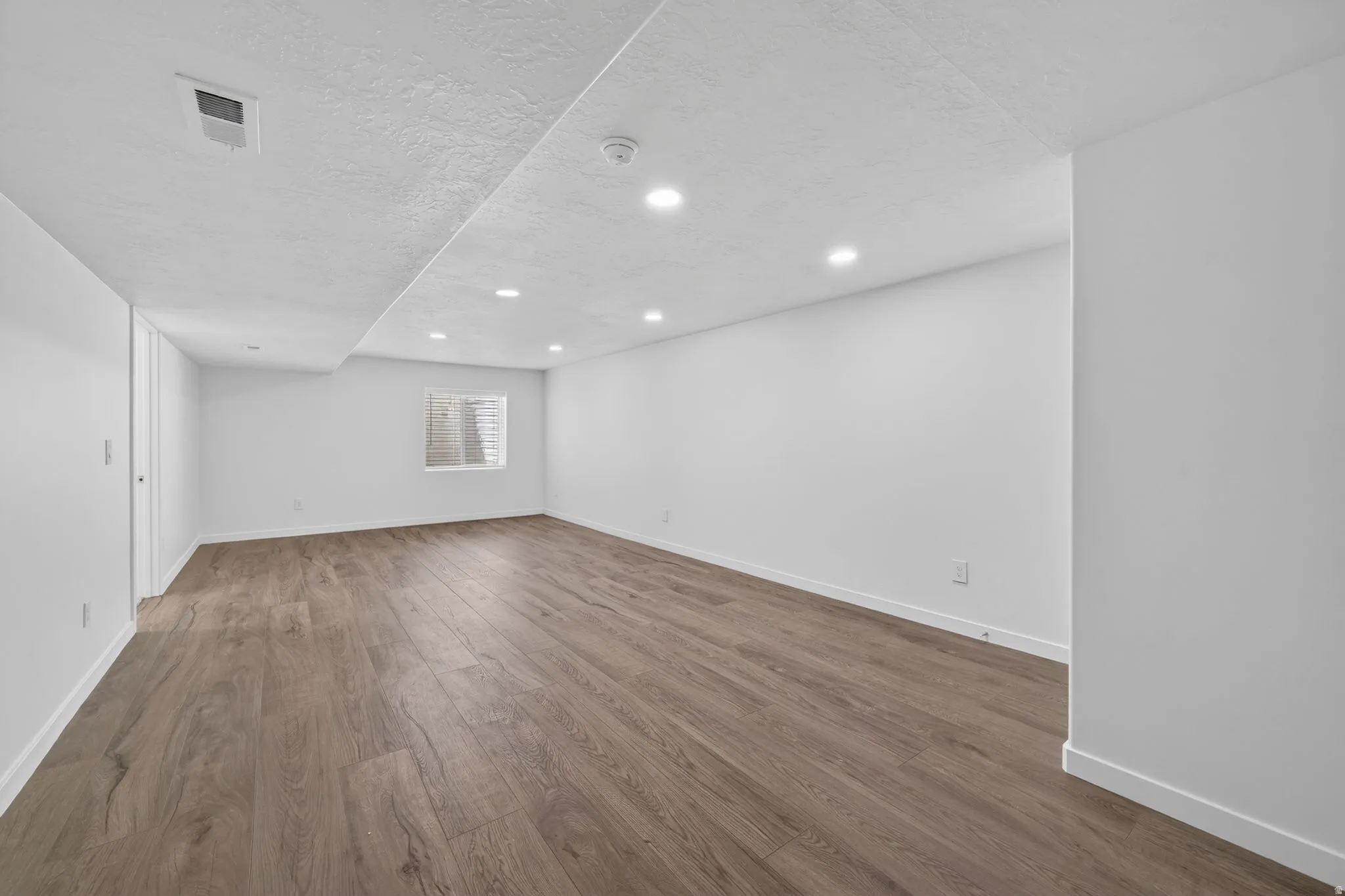 Expansive room featuring wood-finish flooring, recessed lighting, a window with horizontal blinds, and a white color palette