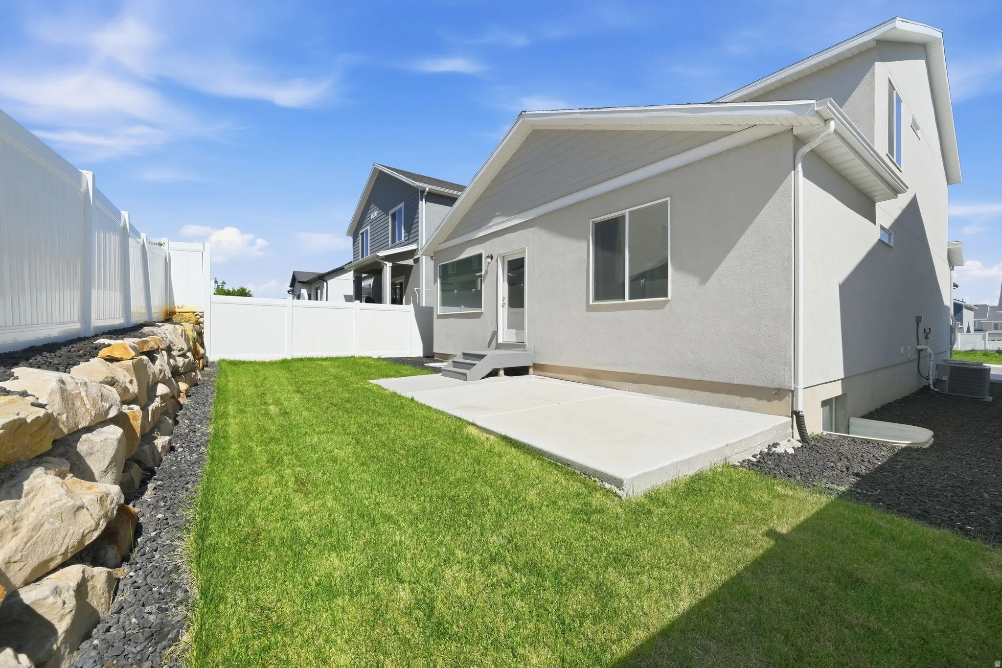 Manicured lawn with a concrete patio, rock retaining wall, and white vinyl fencing
