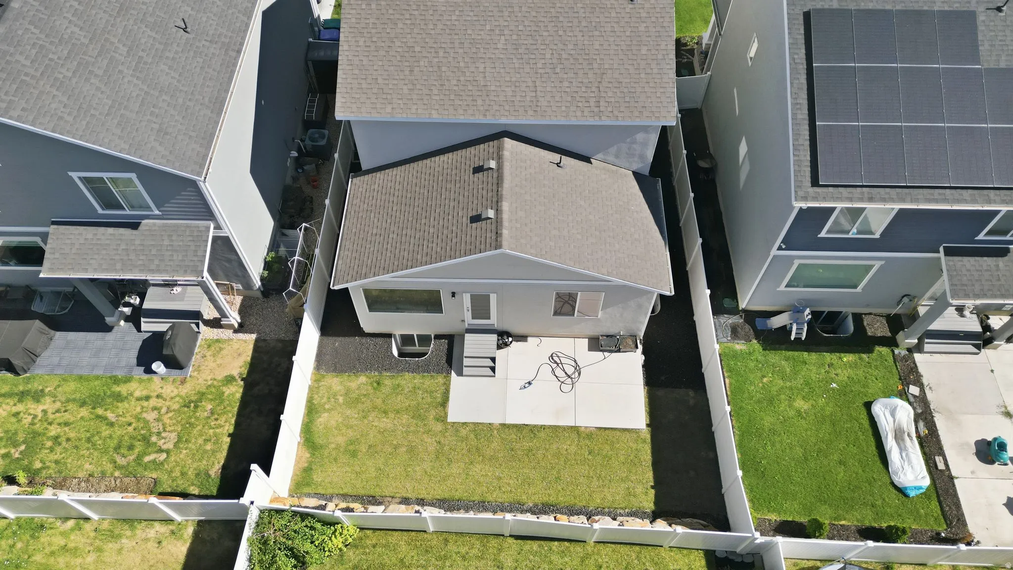 Expansive backyard featuring a concrete patio, a section of manicured lawn, a white PVC fence, and a basement window well