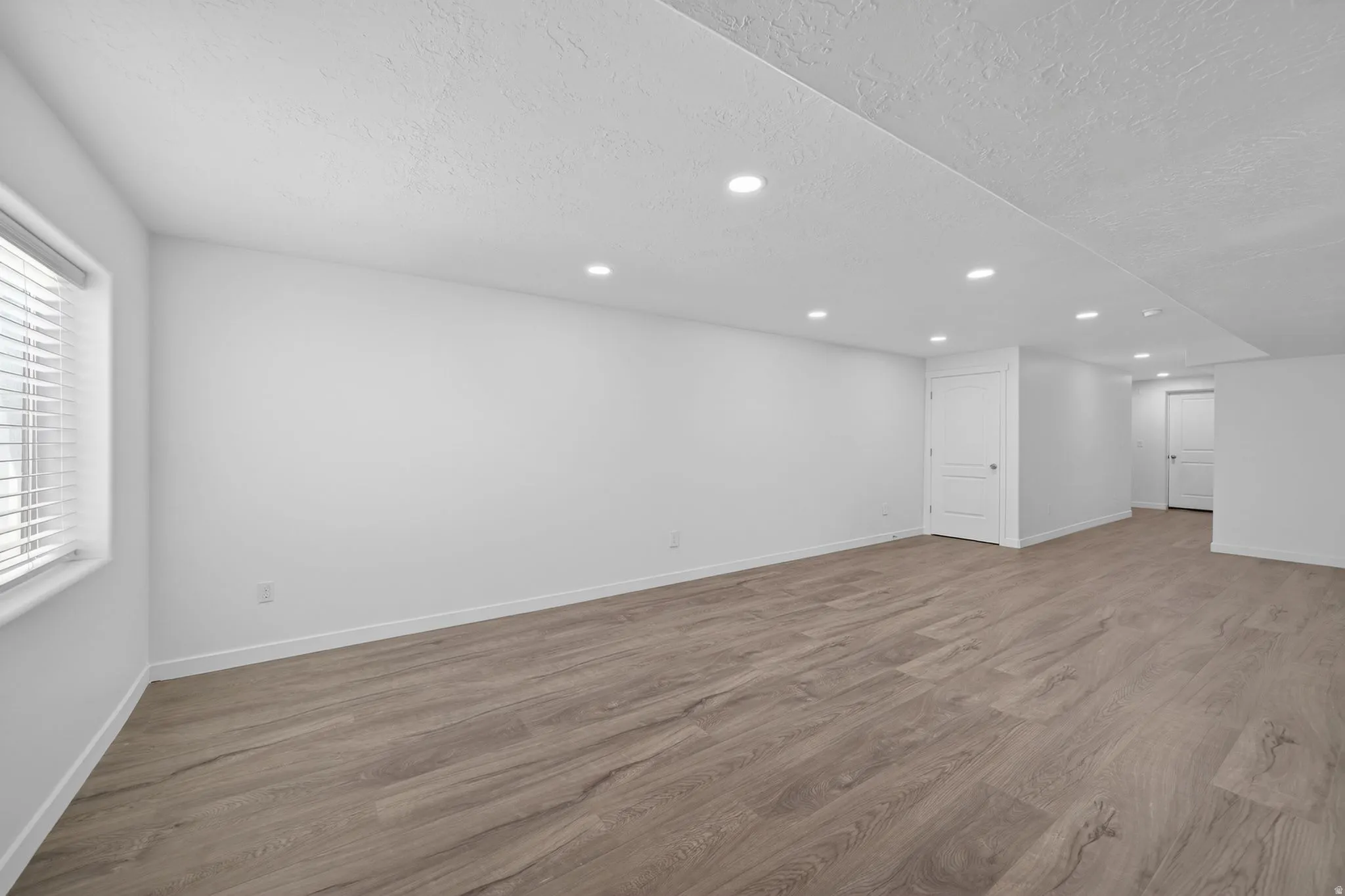 Spacious room featuring wood-finish flooring, white walls, recessed ceiling lighting, and a window with blinds