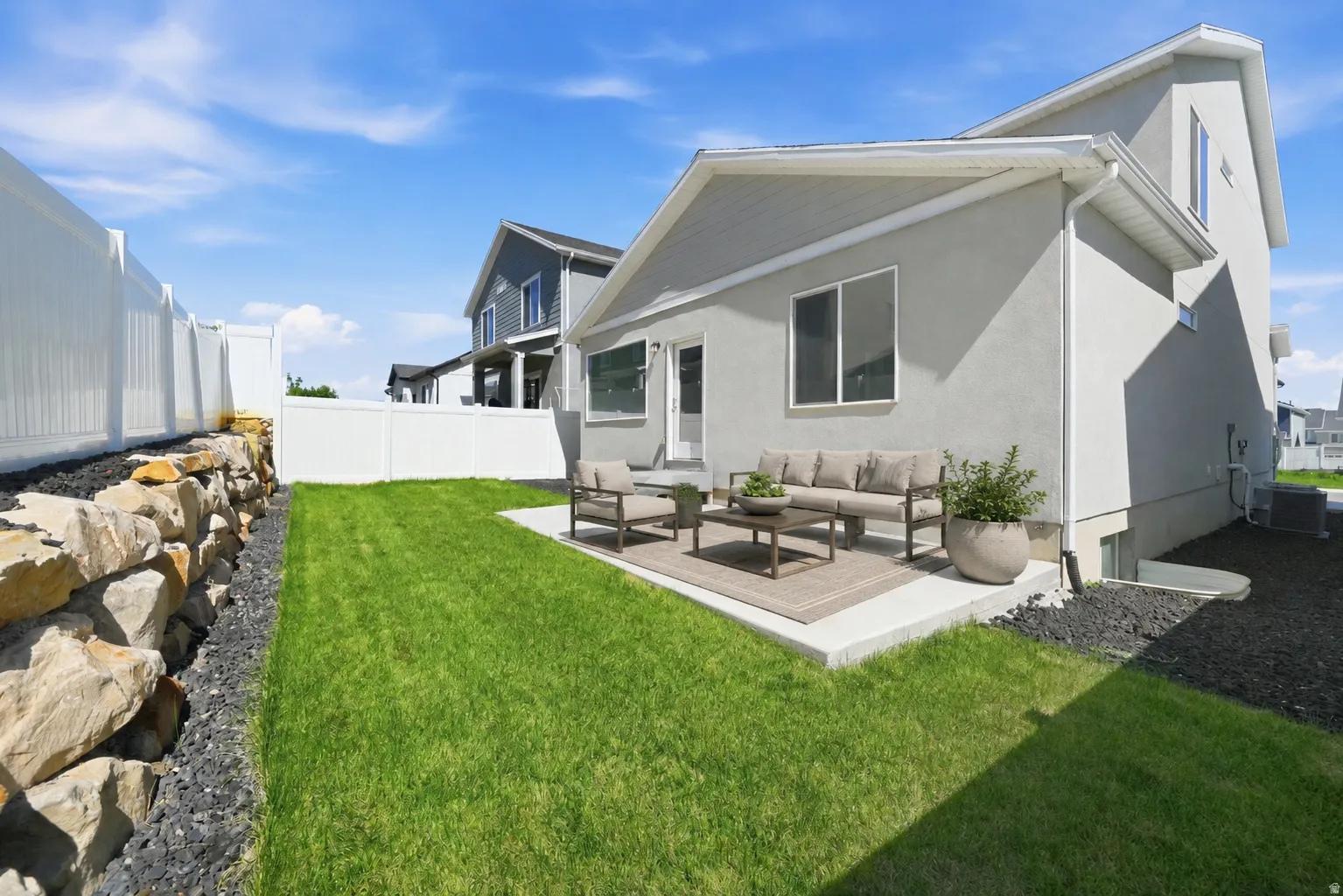 Manicured backyard featuring a concrete patio, lush green lawn, and a natural stone retaining wall with dark rock ground cover