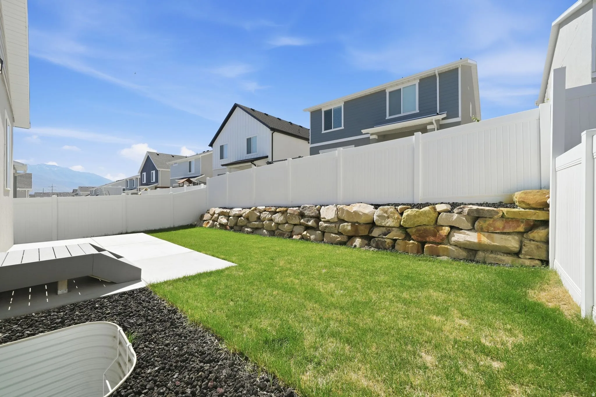 Fenced backyard featuring a manicured lawn, a stone retaining wall, and a concrete patio with an attached deck