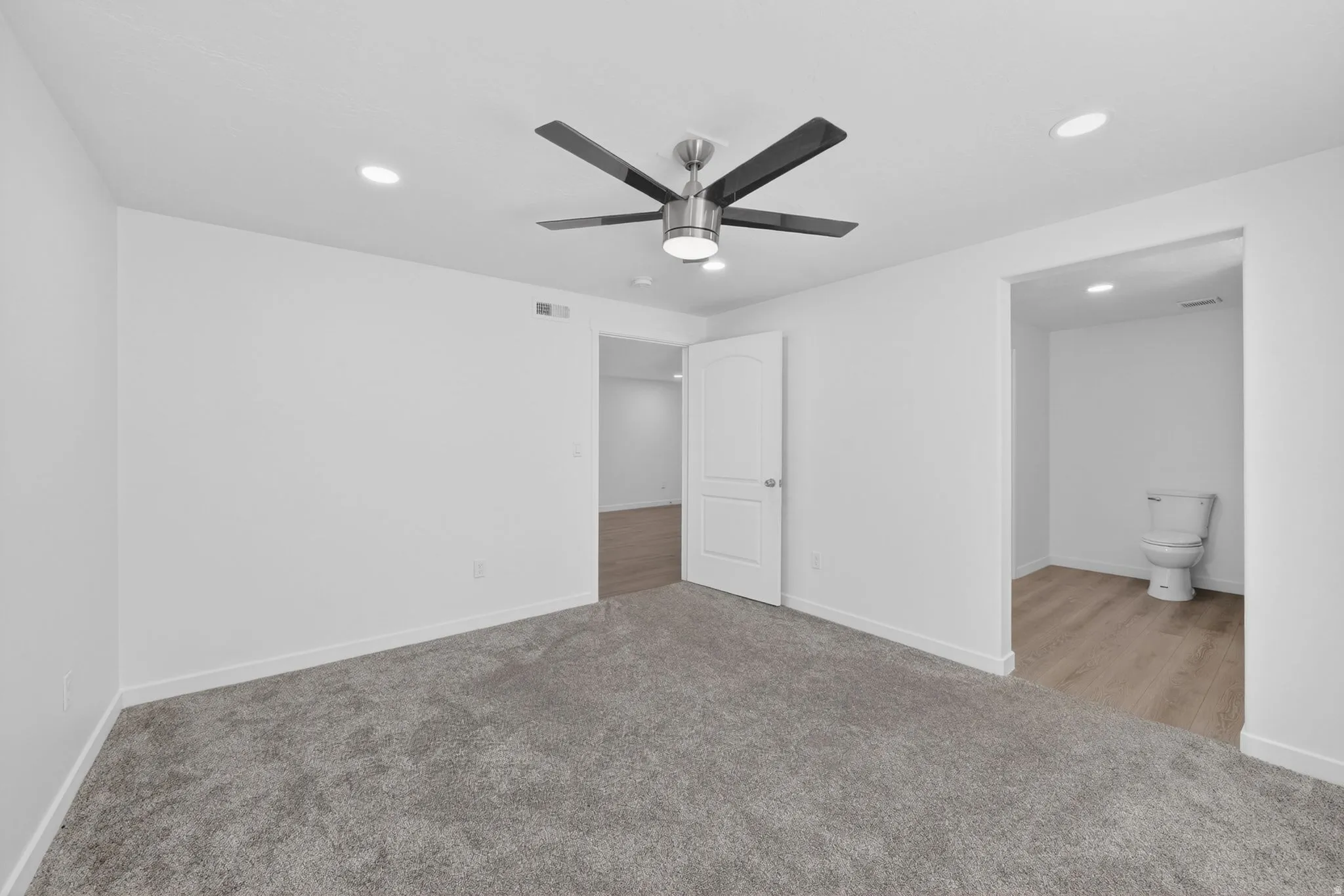 Carpeted room featuring a ceiling fan with integrated lighting, recessed ceiling lights, a white paneled door, and an entryway to a space with wood-finish flooring and a toilet