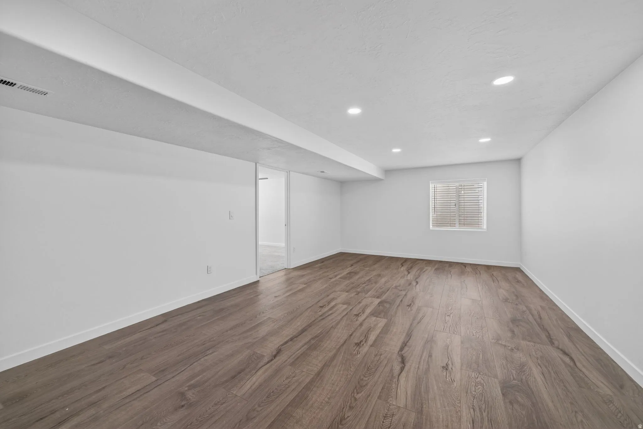 Spacious room with wood-finish flooring, recessed lighting, a single window with blinds, and white baseboards