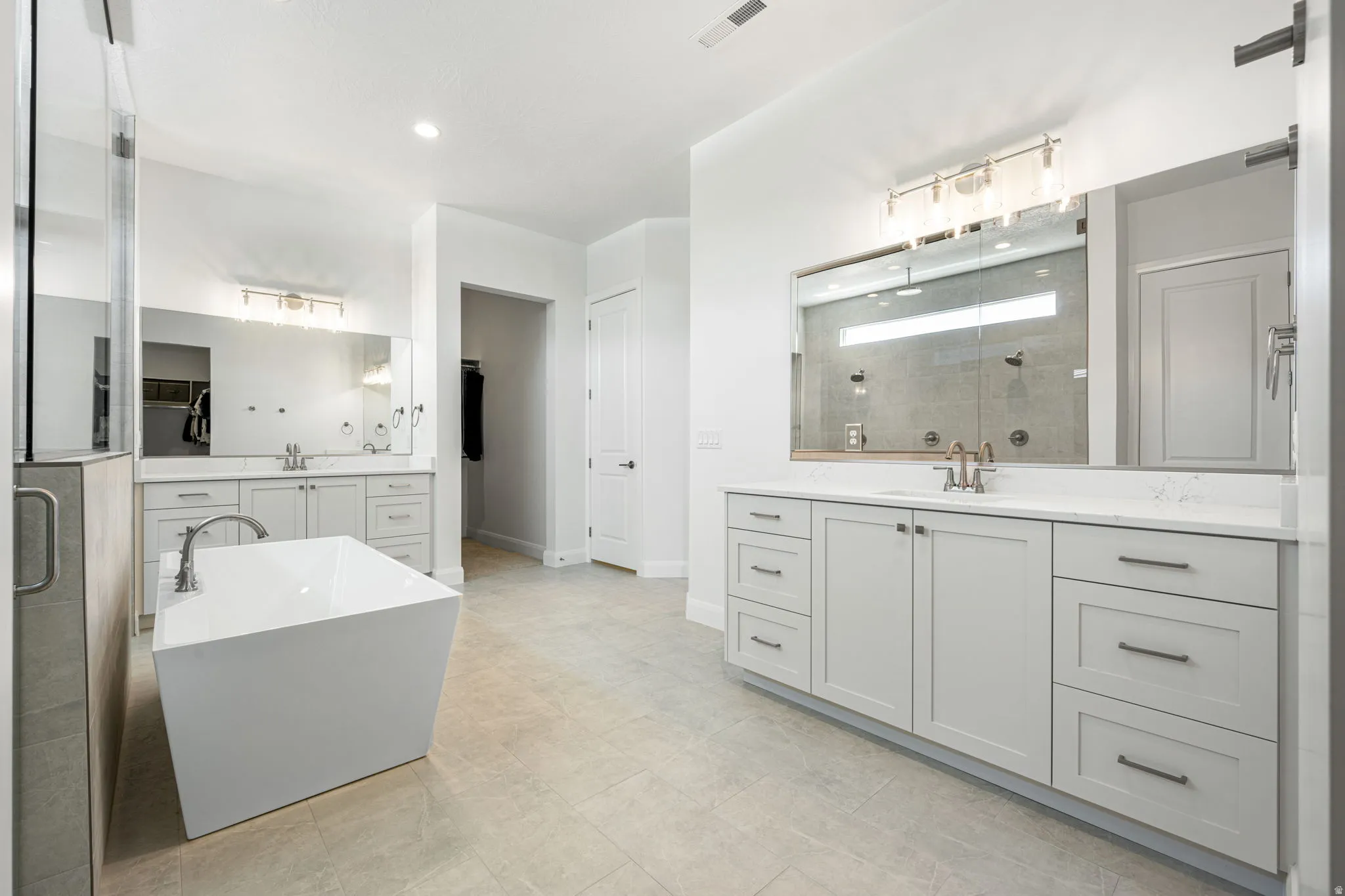 Spacious bathroom featuring a freestanding soaking tub, dual vanities with light cabinetry and solid-surface countertops, a large walk-in shower with glass enclosure, and light tile flooring