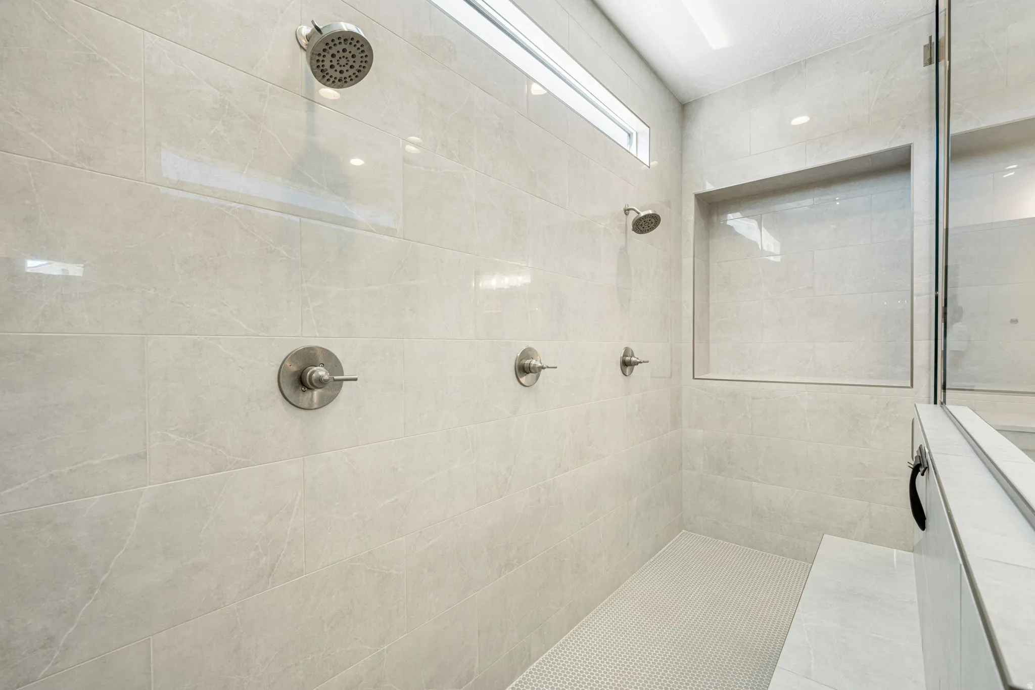Spacious shower featuring light-toned large format wall tiles, a mosaic tile floor, multiple shower heads, a built-in bench, and a convenient recessed shelf