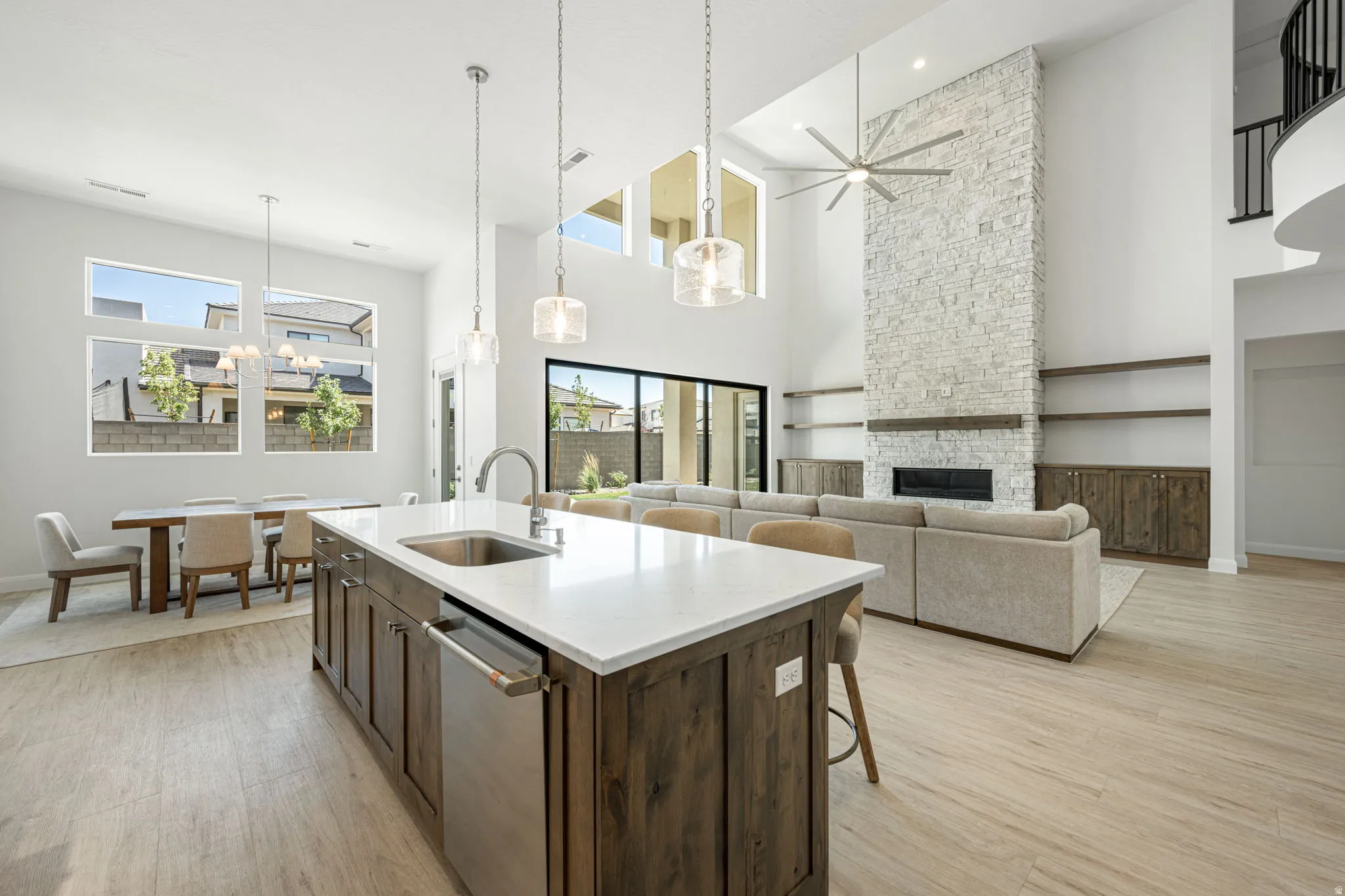Open-concept living space with wood-finish flooring, a kitchen island featuring a stainless steel sink and appliance, a stacked stone fireplace with a wood mantle, and high ceilings with upper-level windows