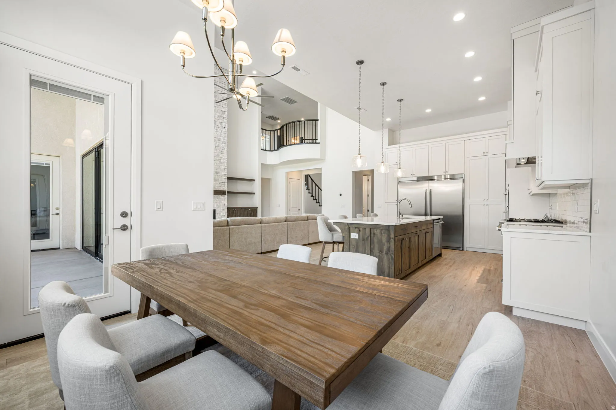 Open-concept living space featuring wood-finish flooring, a contemporary chandelier, a kitchen with white cabinetry and stainless steel appliances, a kitchen island with a light-toned countertop, and a double-height ceiling in the background