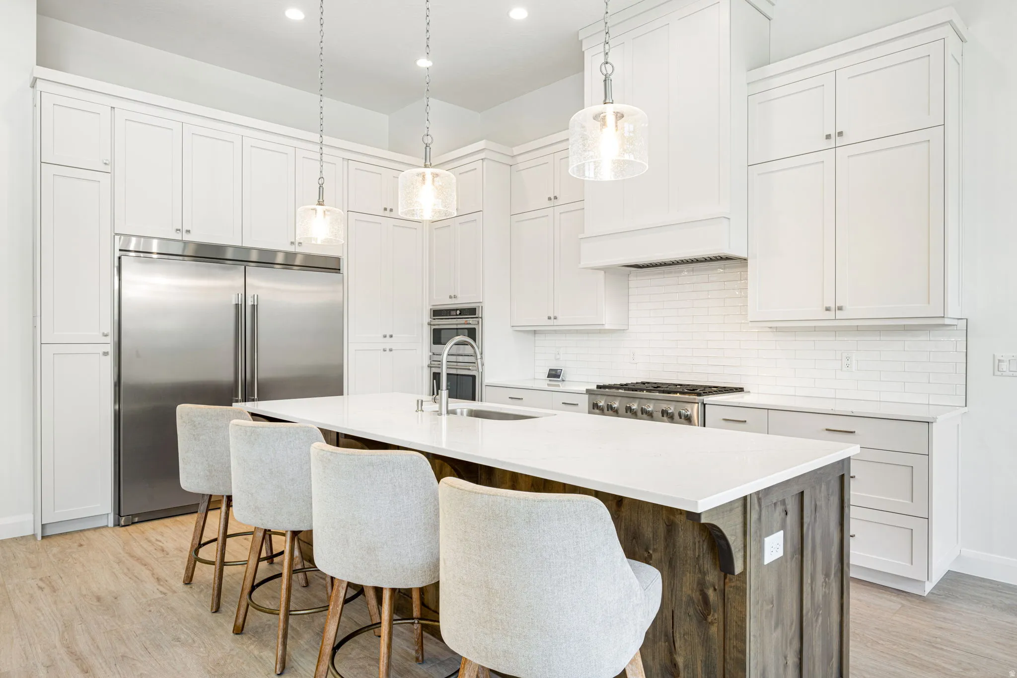 Modern kitchen featuring a large central island with seating for four, wood-finish flooring, white shaker-style cabinetry, stainless steel appliances, and a subway tile backsplash