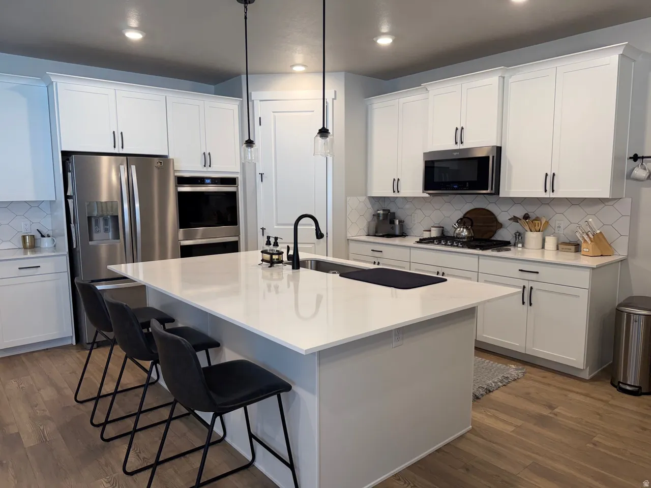 Bright kitchen featuring a substantial island with countertop seating, white shaker-style cabinetry, stainless steel appliances, a white geometric tile backsplash, and wood-finish flooring