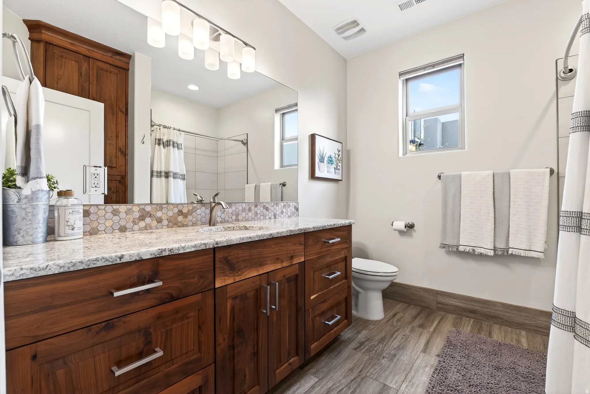 Double vanity with granite countertops, hexagonal tile backsplash, wood-finish cabinetry, and brushed nickel fixtures