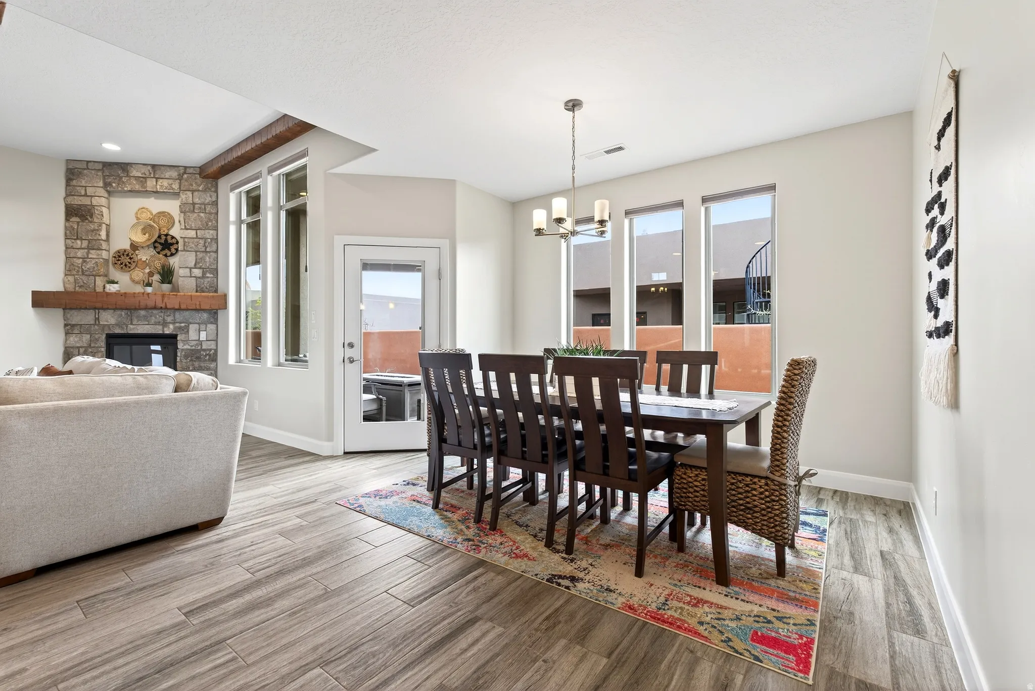 Open-concept dining and living area featuring wood-finish flooring, a stacked stone fireplace with a substantial wood mantel, a clear glass paneled door, and recessed lighting