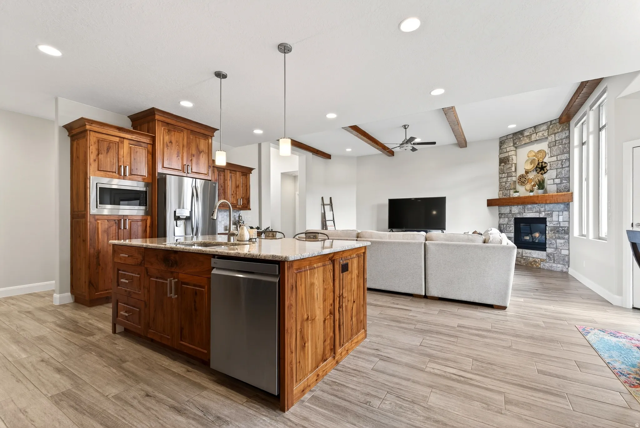 Open-concept living space featuring a spacious kitchen island with integrated sink, stainless steel appliances, wood-finish flooring, exposed ceiling beams, and a stone-clad fireplace