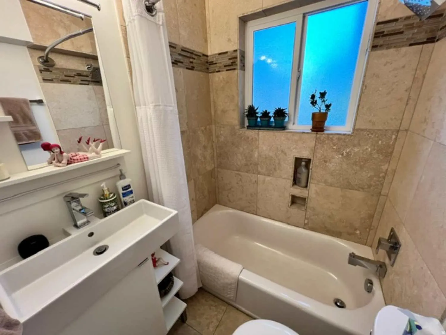 Bathroom featuring a white rectangular vessel sink, floor-to-ceiling tile work with a decorative mosaic accent band, a built-in tub with chrome fixtures, a frosted glass window, and a recessed wall niche