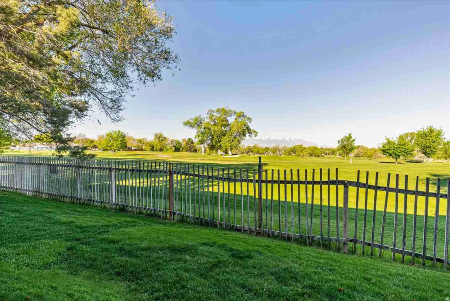 Expansive green lawn bordered by a wood fence, overlooking a broad grassy area with distant trees and mountains