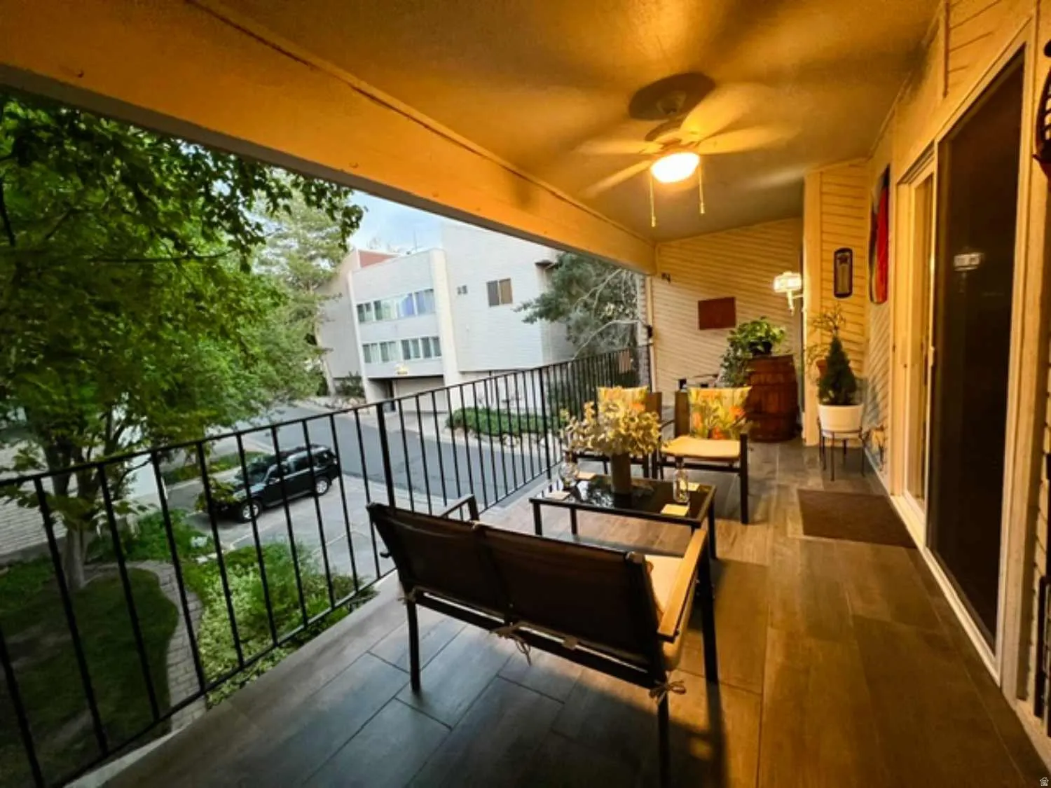 Covered balcony with wood-finish tile flooring, ceiling fan with integrated lighting, and black metal railing