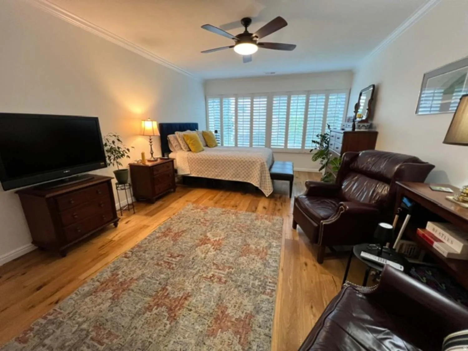 Spacious bedroom featuring wood-finish flooring, a ceiling fan with integrated lighting, crown molding, and plantation shutters
