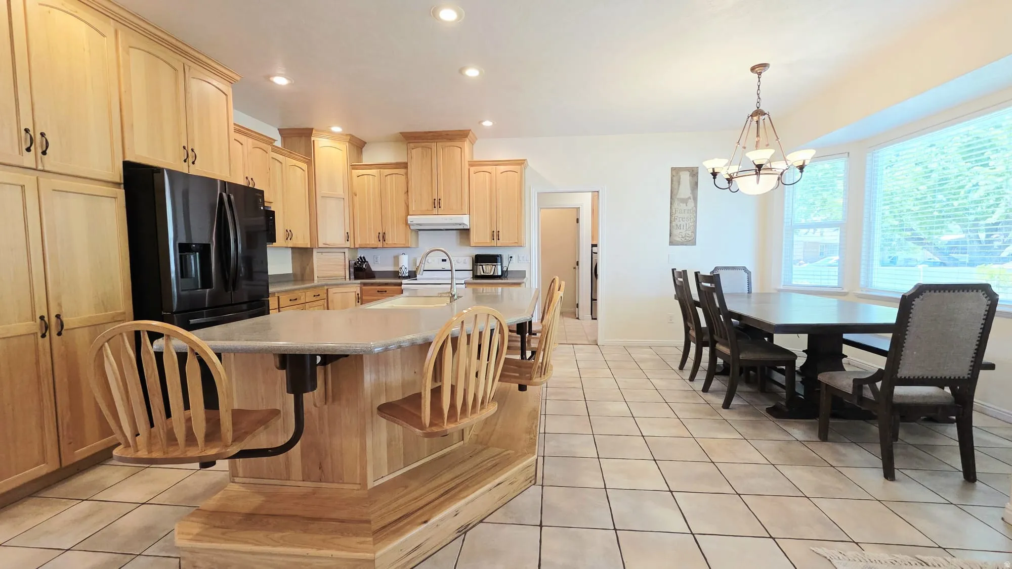 Kitchen and dining area featuring light wood cabinetry, a central island with a light-toned countertop, black appliances, tile flooring, and ample natural light from multiple windows