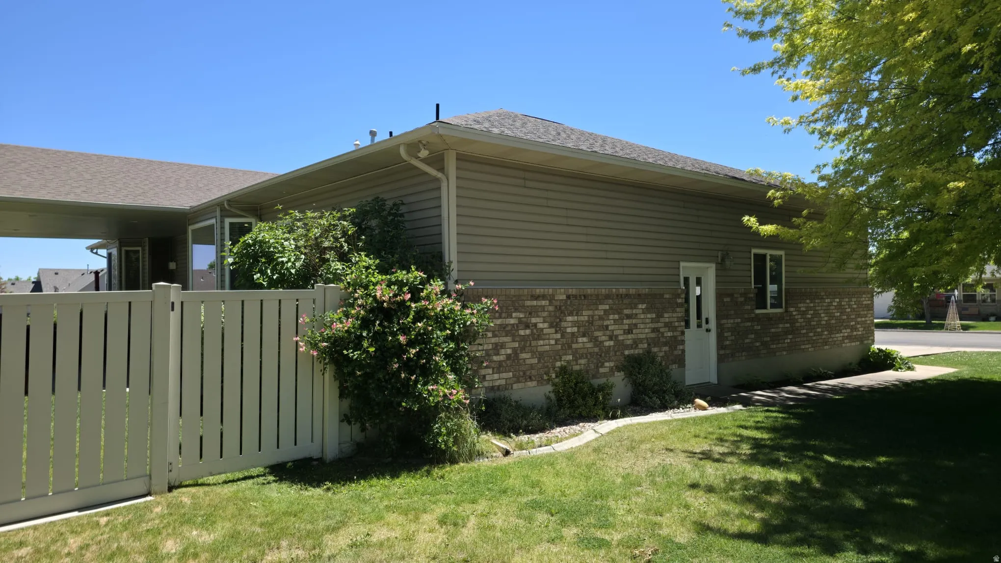 Exterior featuring beige horizontal siding, brick veneer, a white privacy fence, and mature landscaping