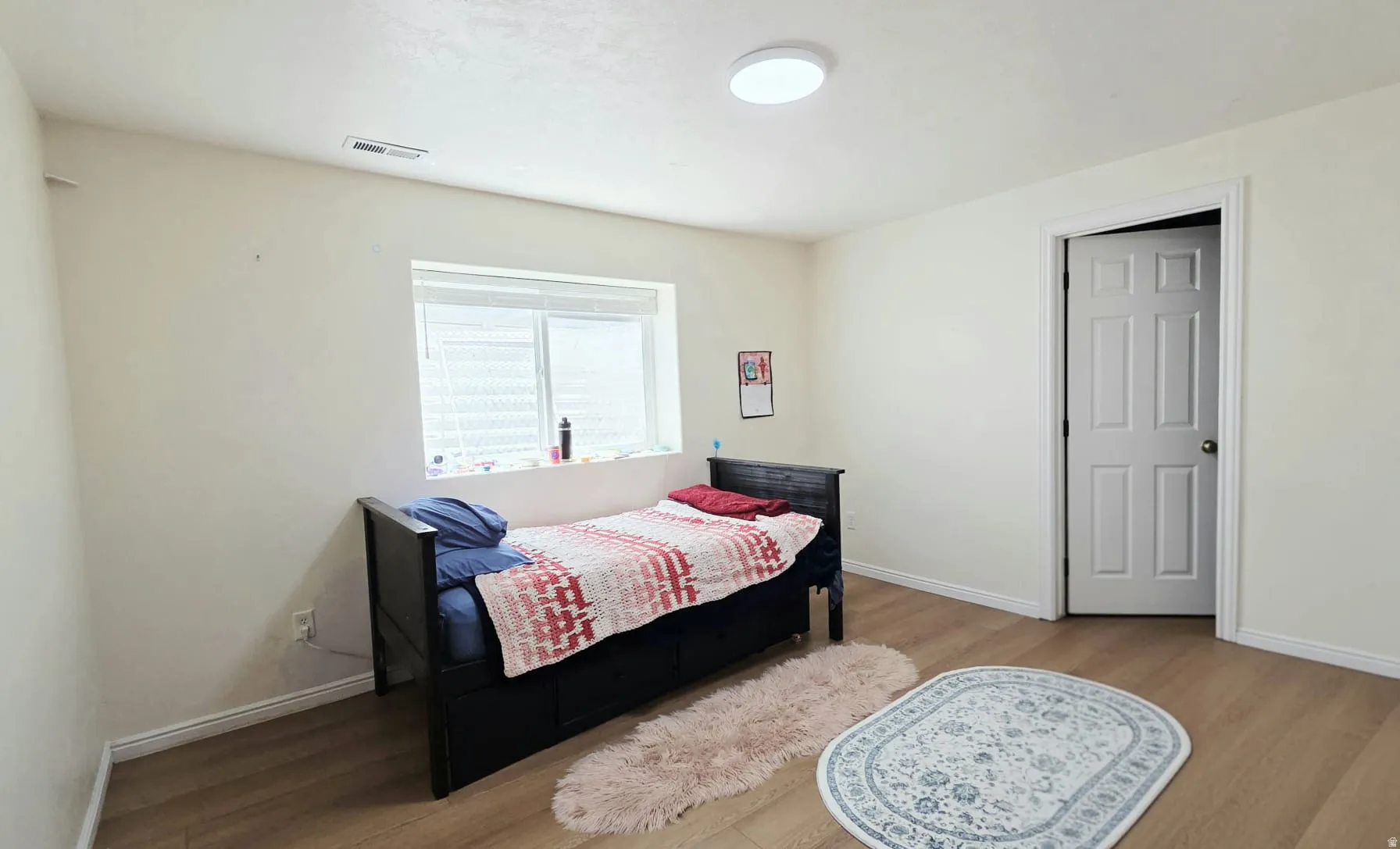 Room featuring light-toned walls, wood-finish flooring, a flush-mount ceiling light, and a window with blinds