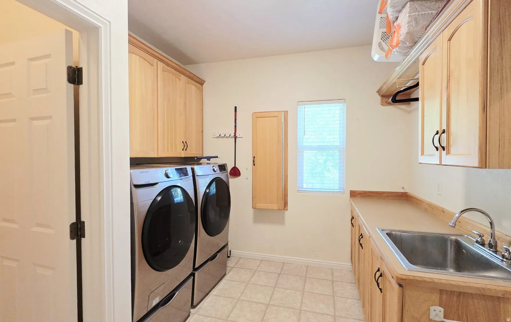 Functional utility room featuring extensive light wood cabinetry, integrated stainless steel sink, tiled flooring, and a window with blinds