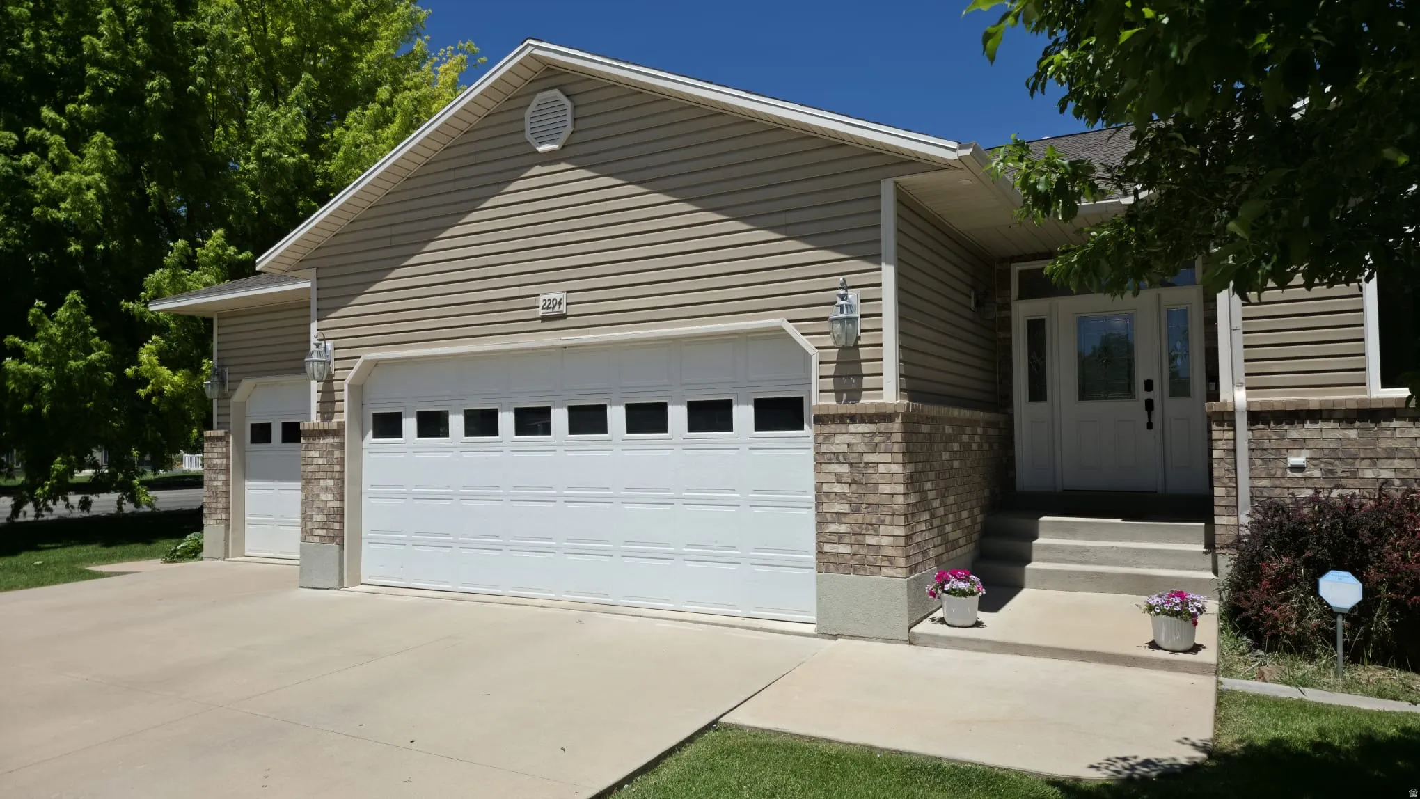 Exterior featuring tan siding, brick accents, a two-car garage with window panels, and a concrete driveway