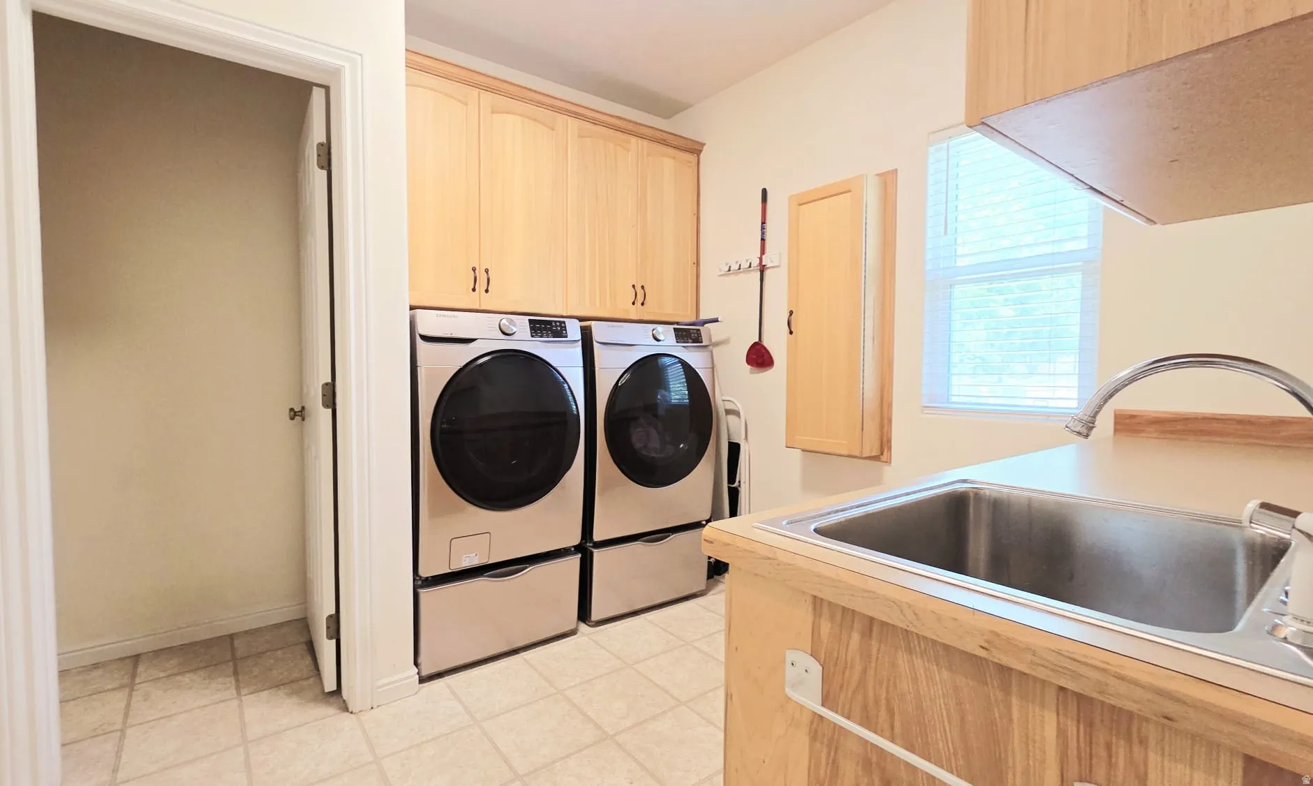 Dedicated laundry room featuring light wood-finish cabinetry, a utility sink with a high-arc faucet, a window with blinds, and tiled flooring