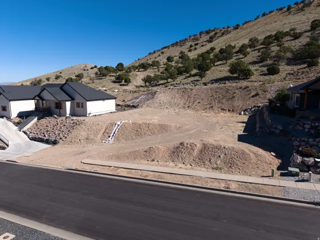 Expansive lot with graded terrain, bordered by a paved road and a concrete sidewalk, featuring a natural hillside backdrop