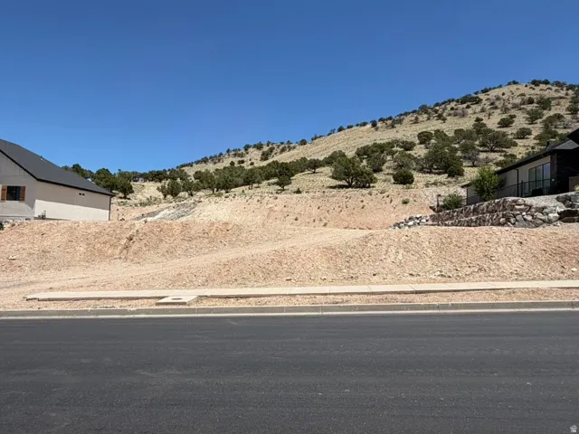 Facing North.  Graded lot featuring a paved road frontage, a concrete sidewalk, and a natural hillside backdrop with scattered trees.