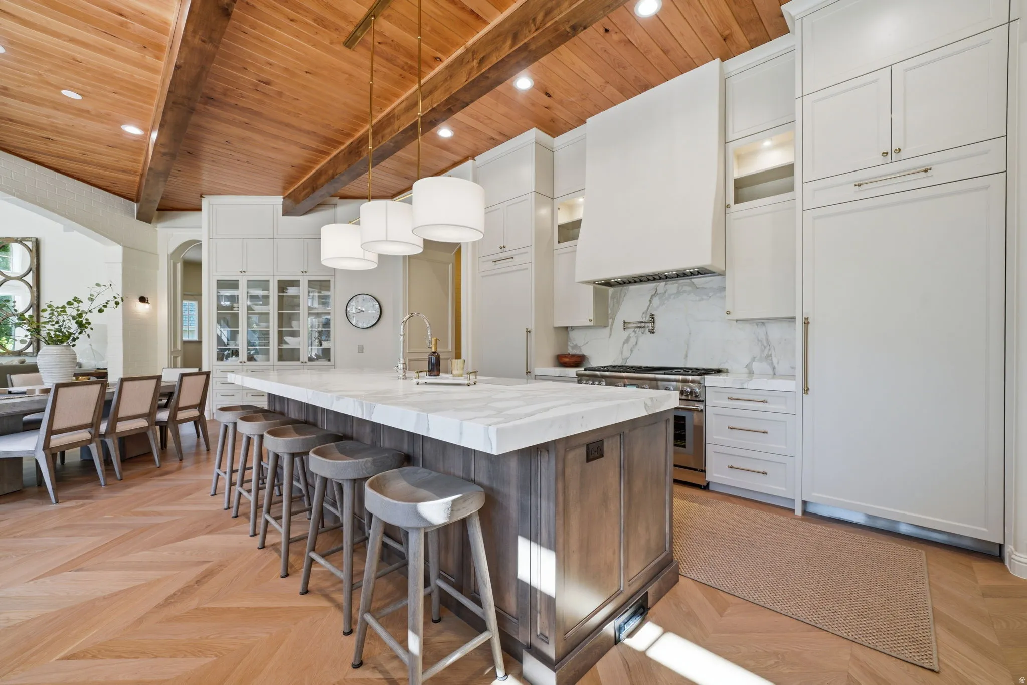 Gourmet kitchen featuring a wood-beam vaulted ceiling, large central island with a waterfall countertop, herringbone wood-finish flooring, white cabinetry, and integrated paneled appliances