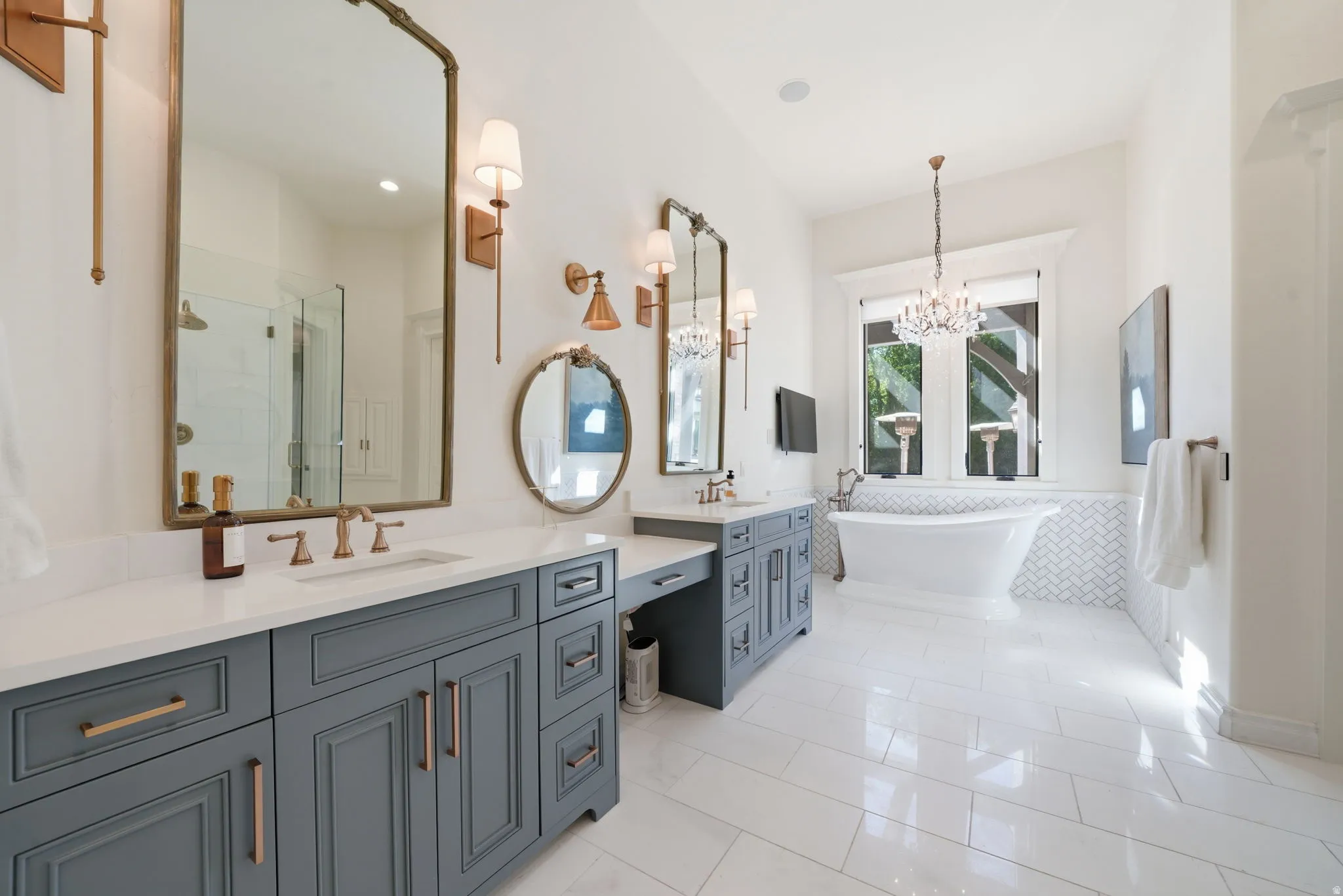 Spacious bathroom featuring a freestanding soaking tub, dual vanity with undermount sinks, separate shower enclosure with glass panel, white tile flooring, and herringbone-patterned wall tile
