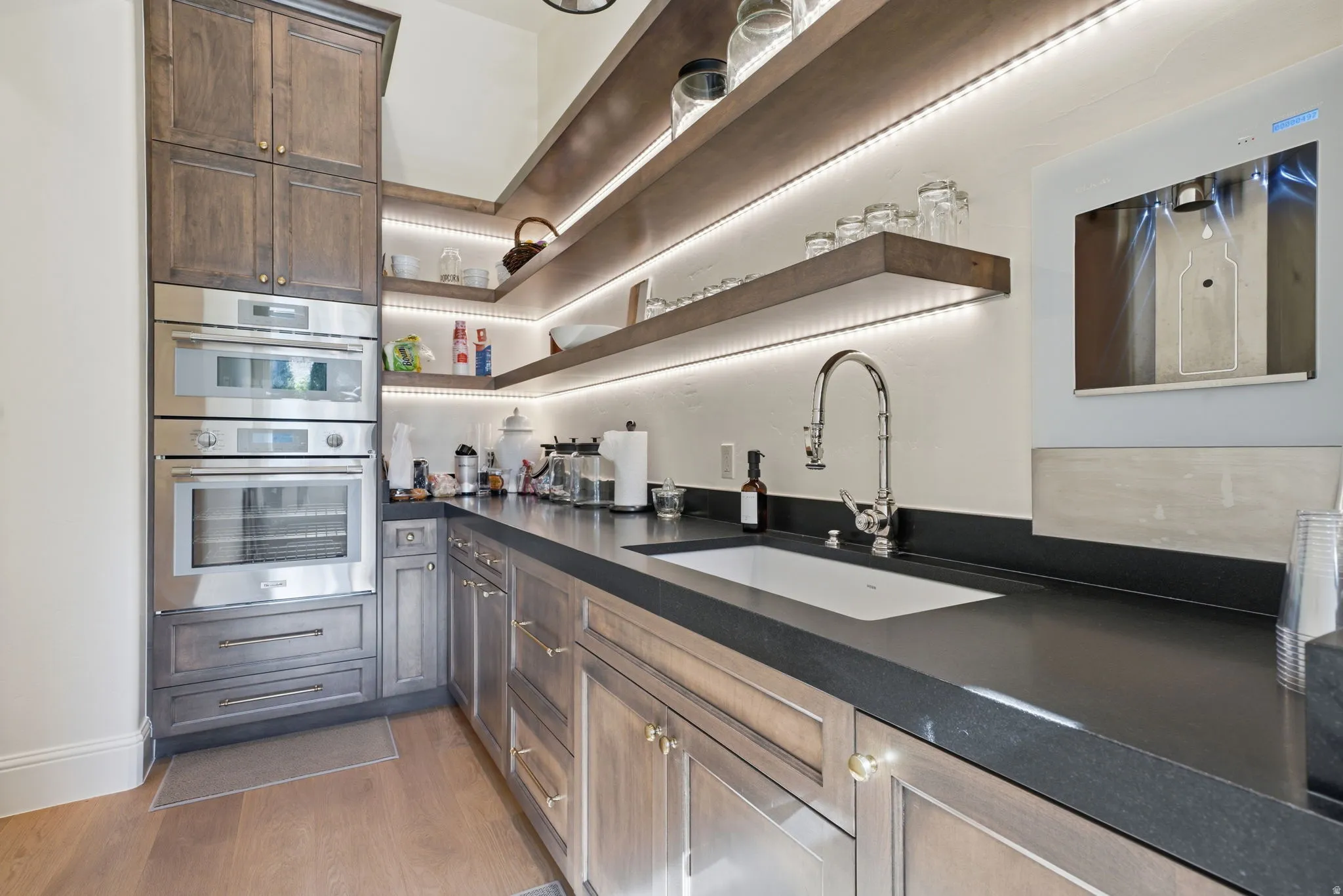 Kitchenette featuring stainless steel double ovens, custom wood cabinetry, under-cabinet lighting, a built-in sink, and dark countertops