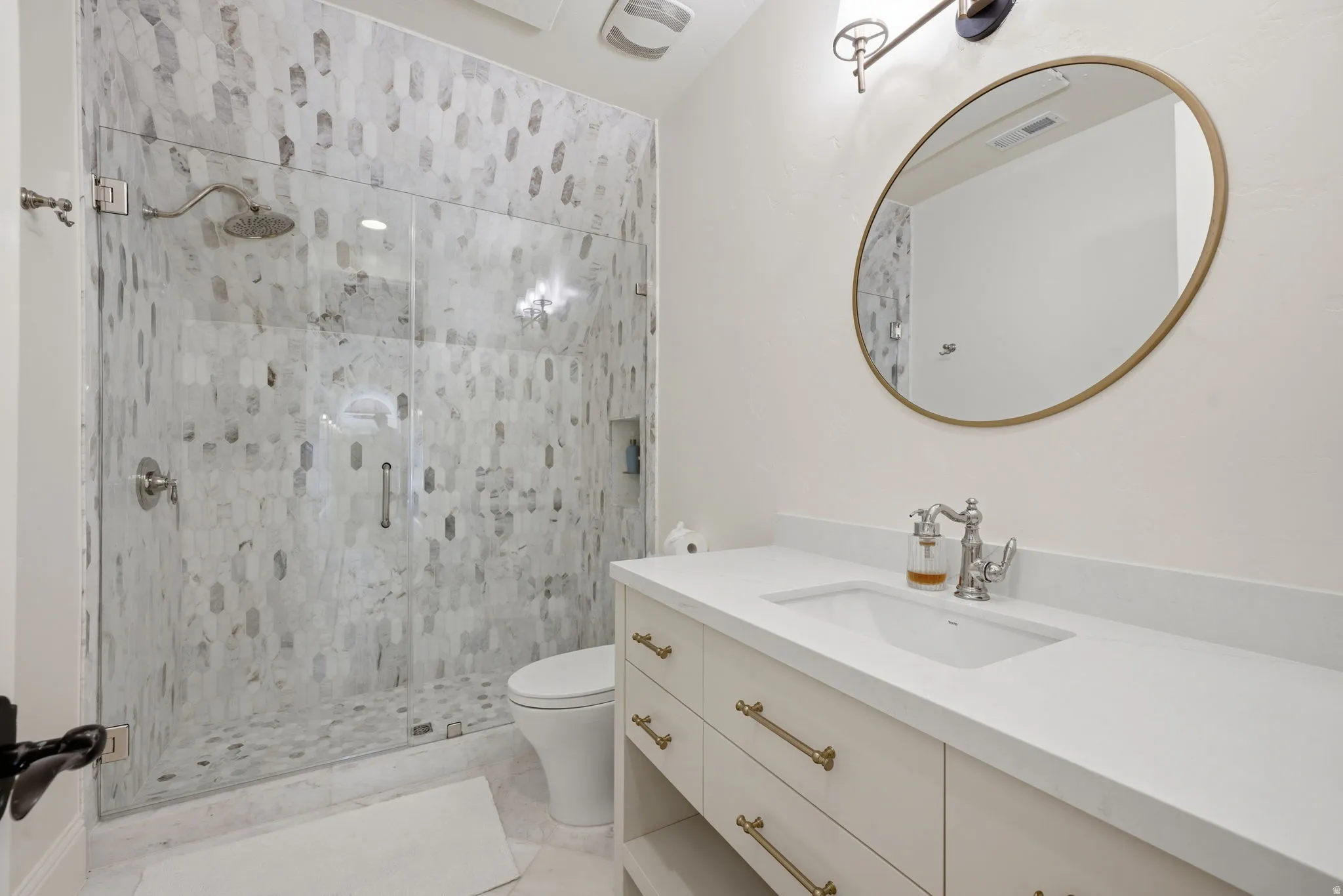 Bathroom featuring a glass-enclosed shower with patterned tile, a single vanity with a white countertop and integrated sink, an oval mirror with a brass frame, and a wall-mounted light fixture